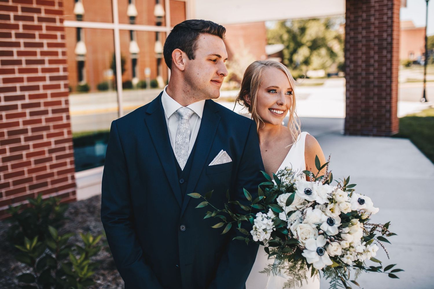 Bride holding white floral bouquet stands with groom in navy suit outside brick building on wedding day.