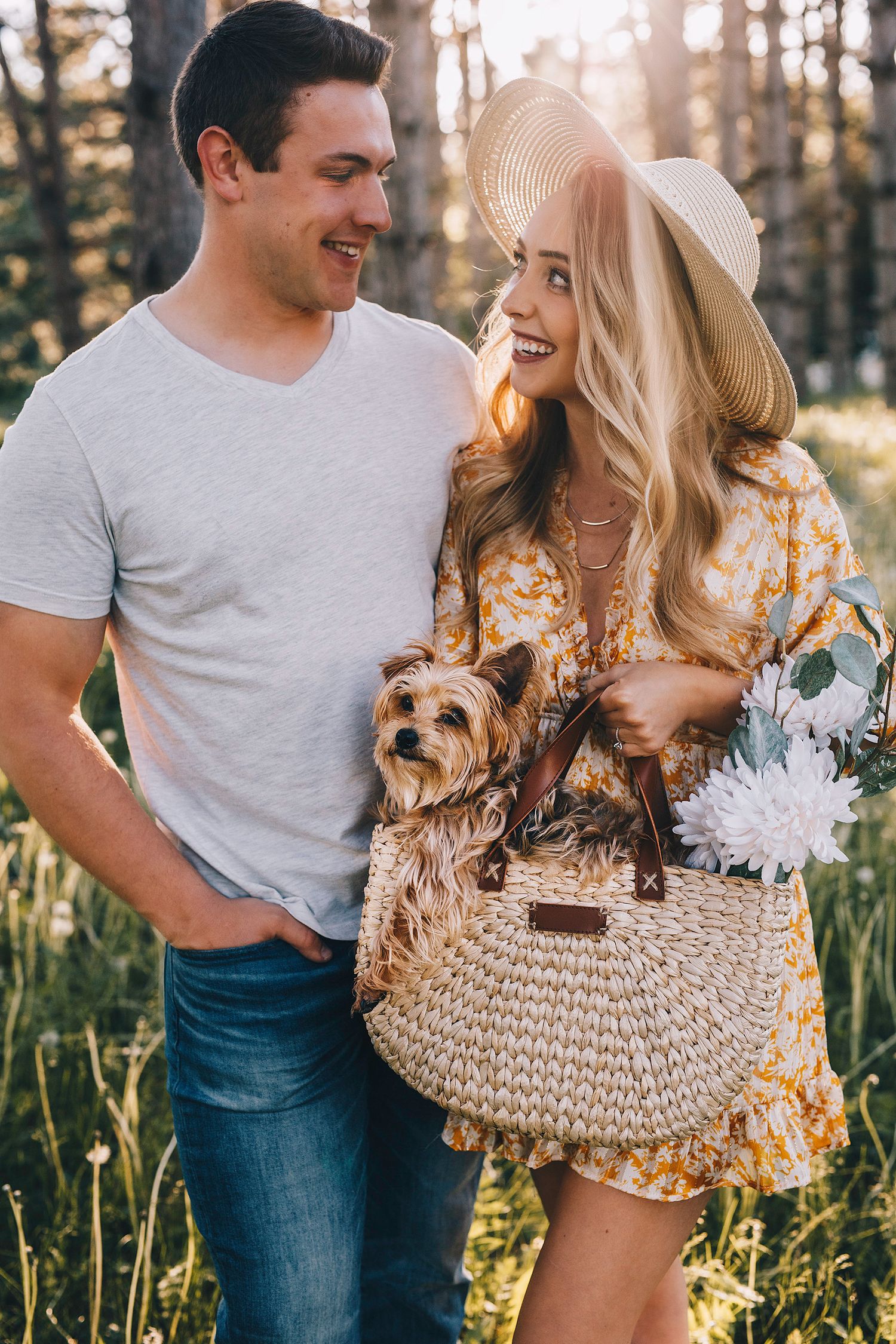 Smiling couple outdoors holding a Yorkshire Terrier in a wicker bag with white flowers.