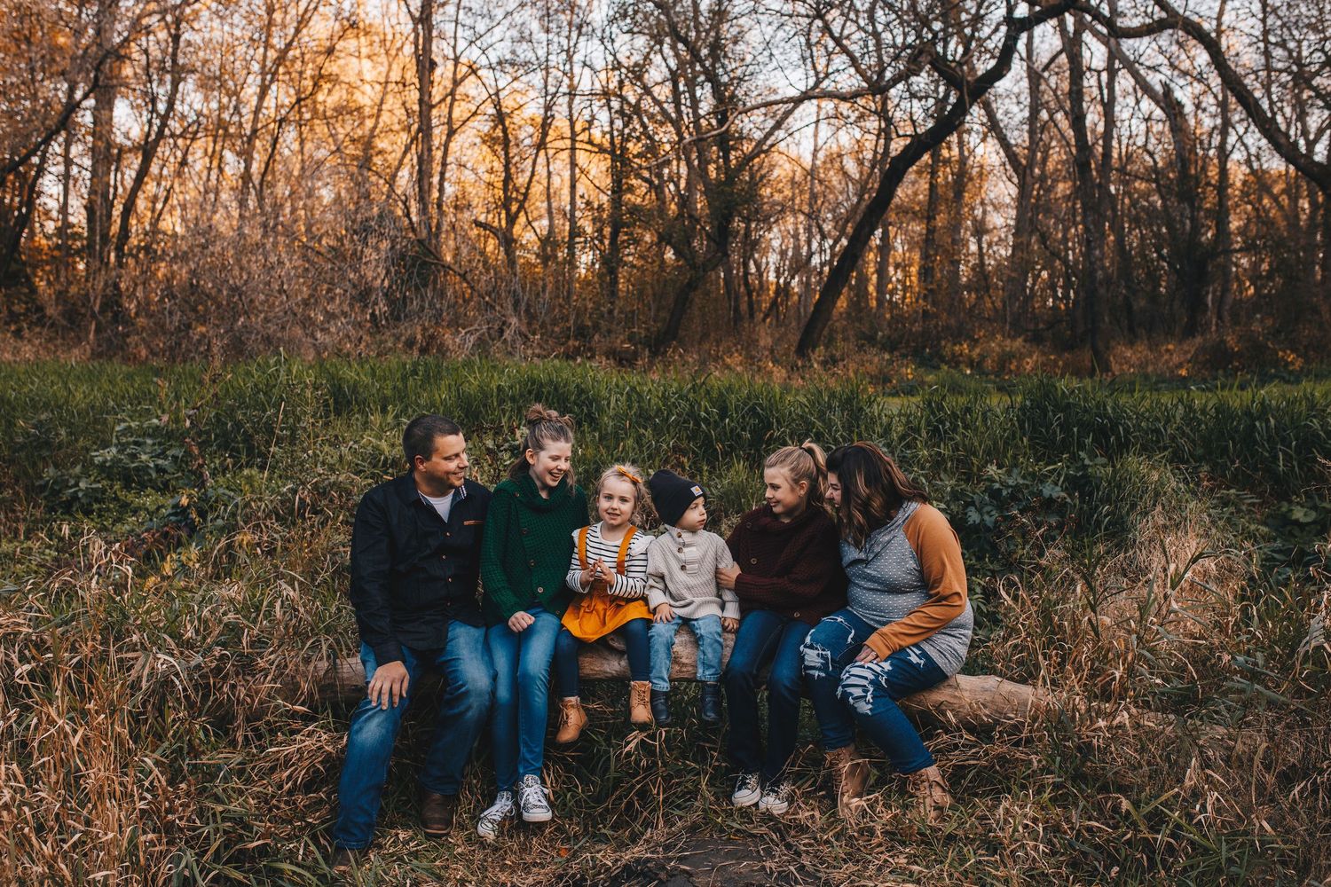 Family of six smiling together on a log in a colorful autumn forest setting.
