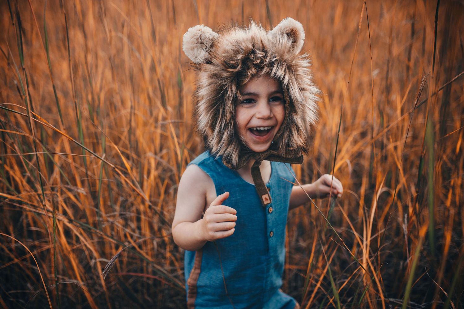 Happy child wearing fuzzy lion hat standing in tall autumn grass, laughing and smiling.