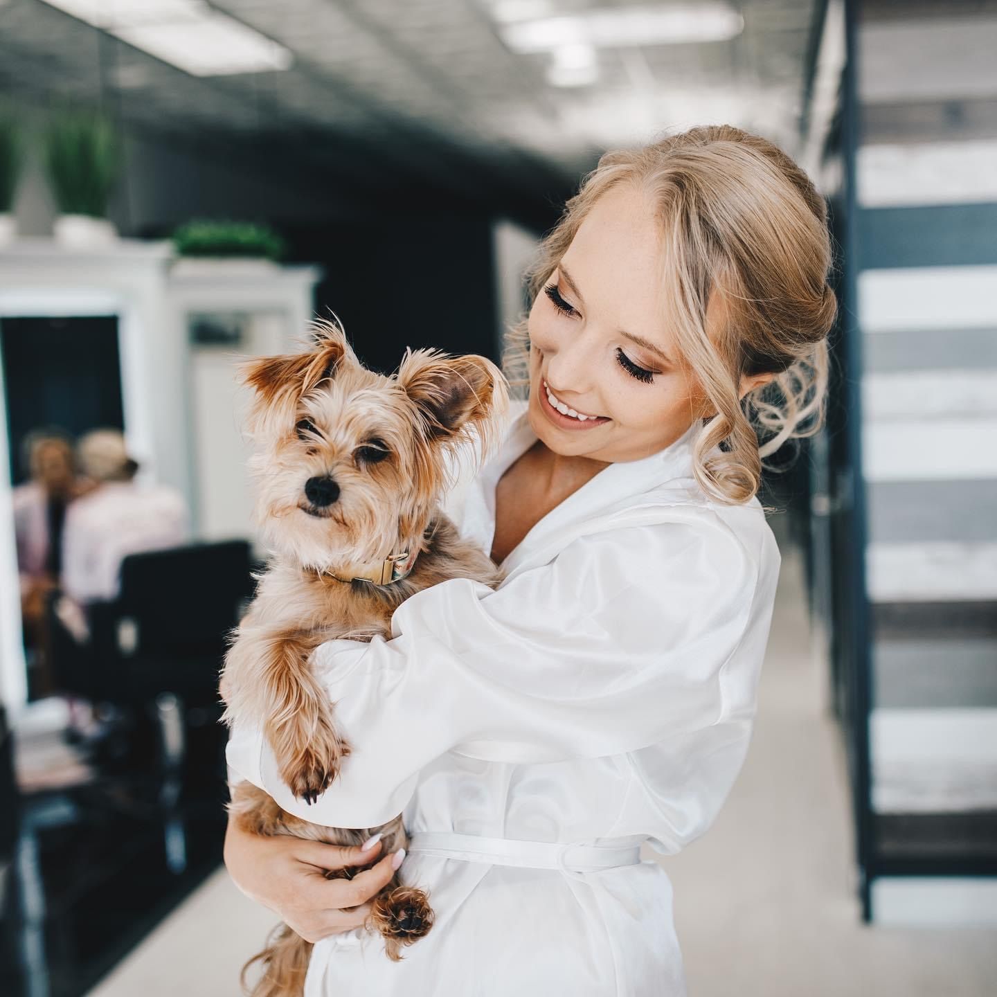 Smiling blonde bride in white robe holding a small Yorkshire Terrier dog indoors.