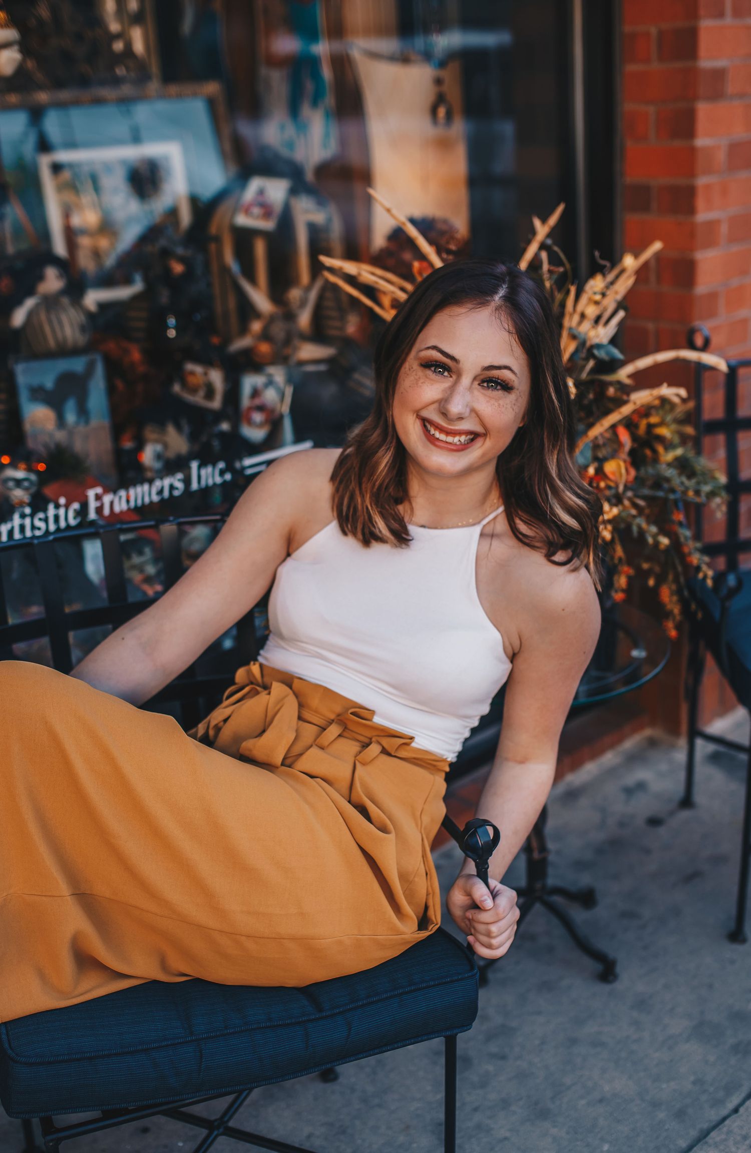 Smiling brunette woman in white top and mustard skirt sitting outside a framing shop.