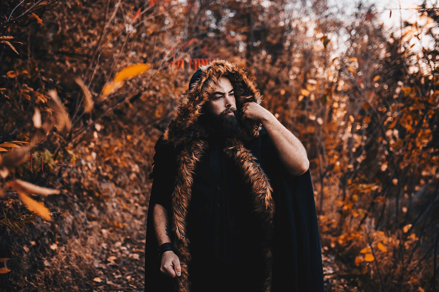 Bearded man in black cape with fur trim standing in autumn forest setting.