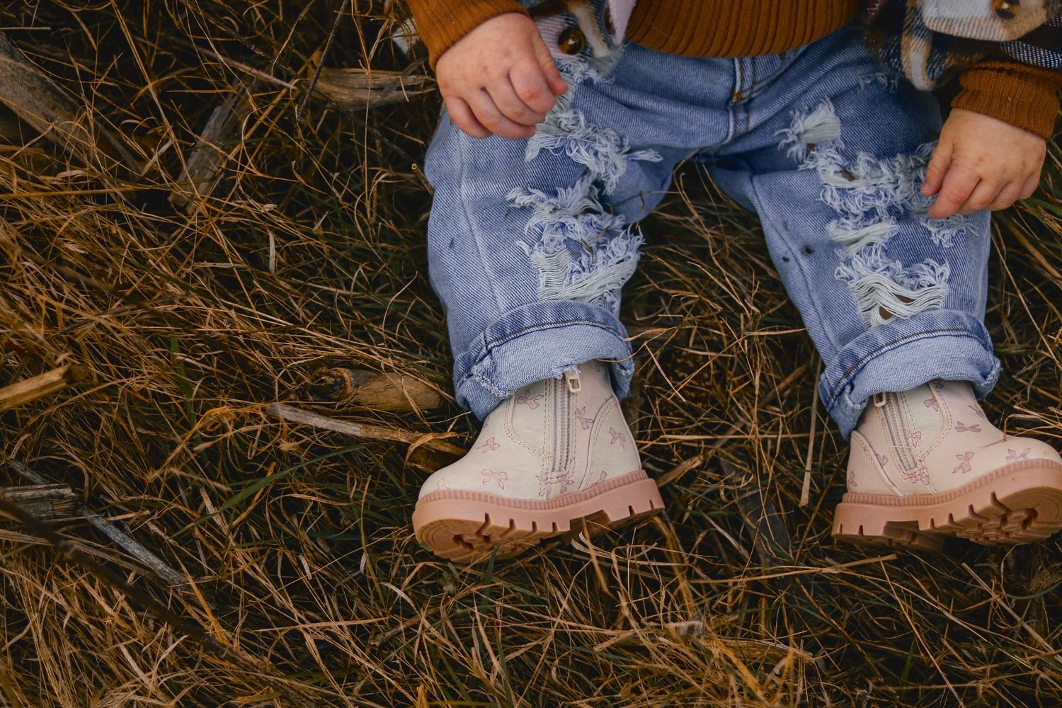 Close-up of toddler in distressed jeans and pink boots sitting on hay.