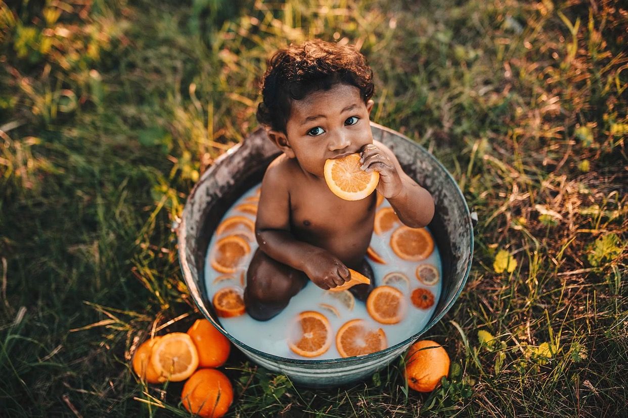 Cute baby sits in metal tub with milk and orange slices on green grass outdoors.