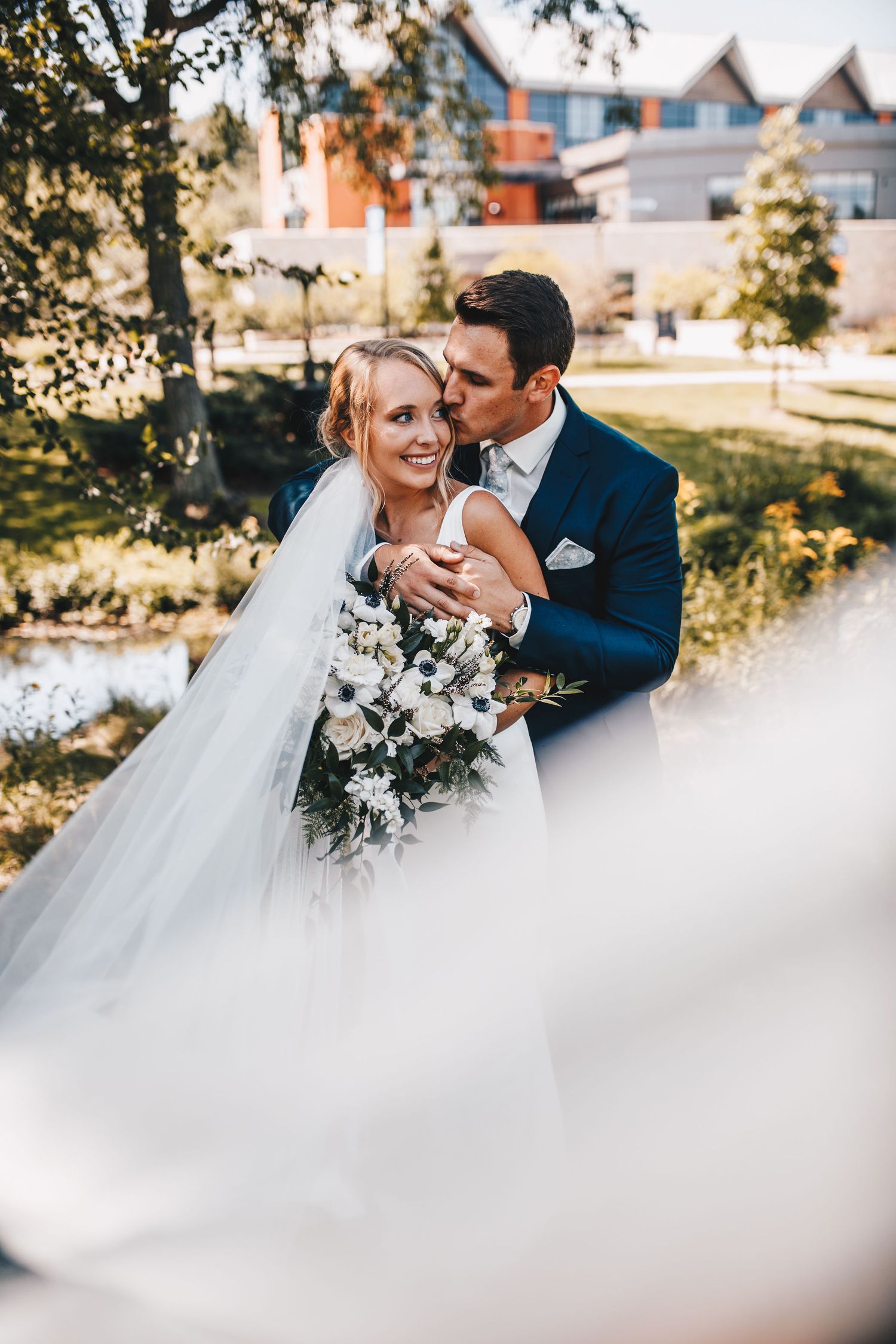 A groom in a navy suit kisses his bride holding a white bouquet in a sunny garden.
