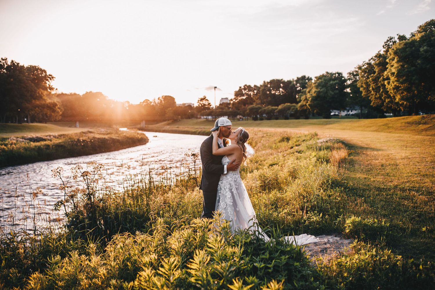 Bride and groom sharing golden hour kiss beside a serene river surrounded by wildflowers.