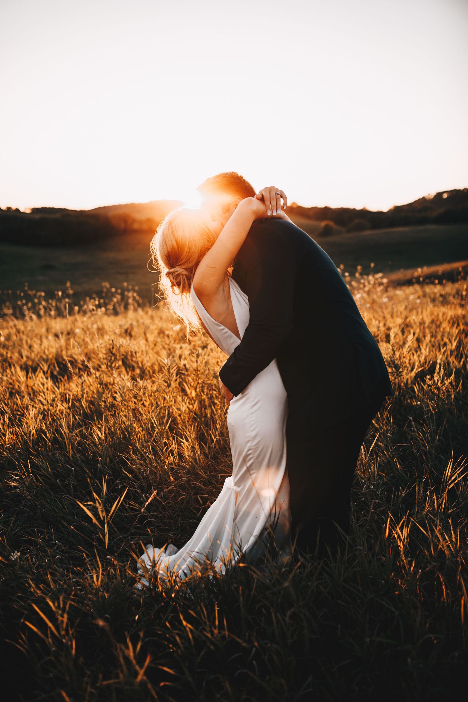 A bride and groom share a romantic kiss in a golden field during a stunning sunset.