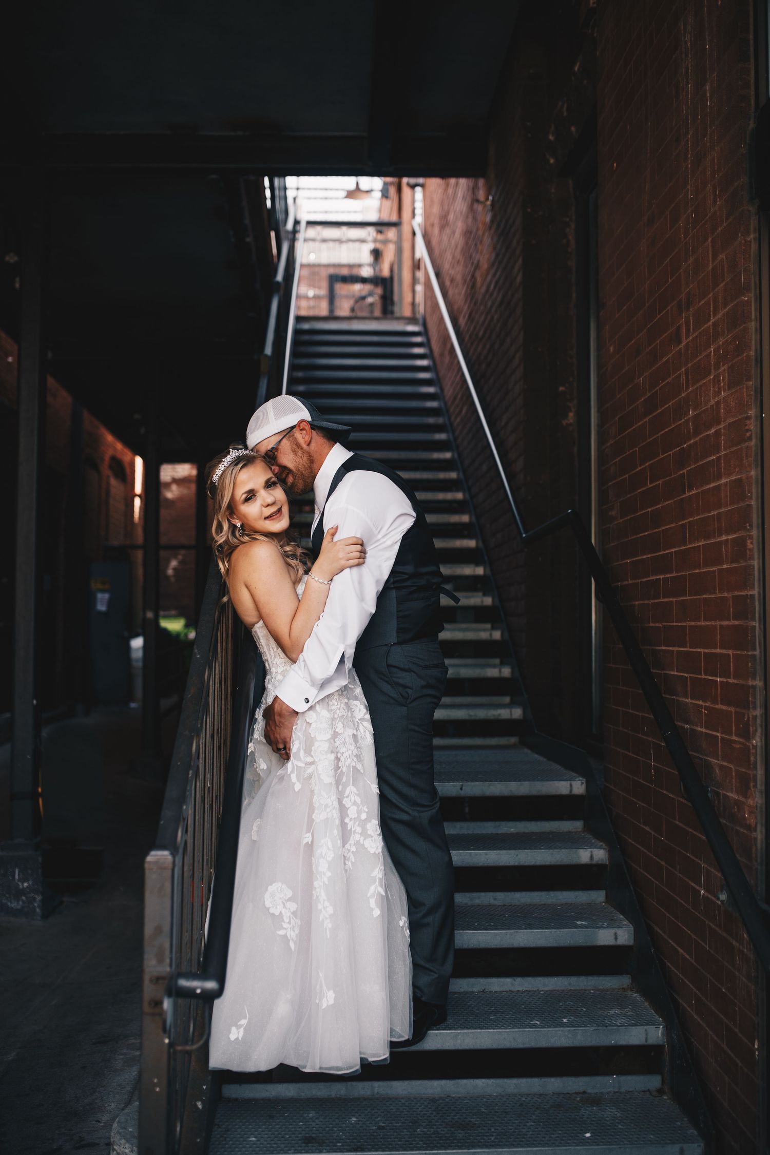 Bride and groom embracing in moody urban alleyway with metal staircase and brick walls.