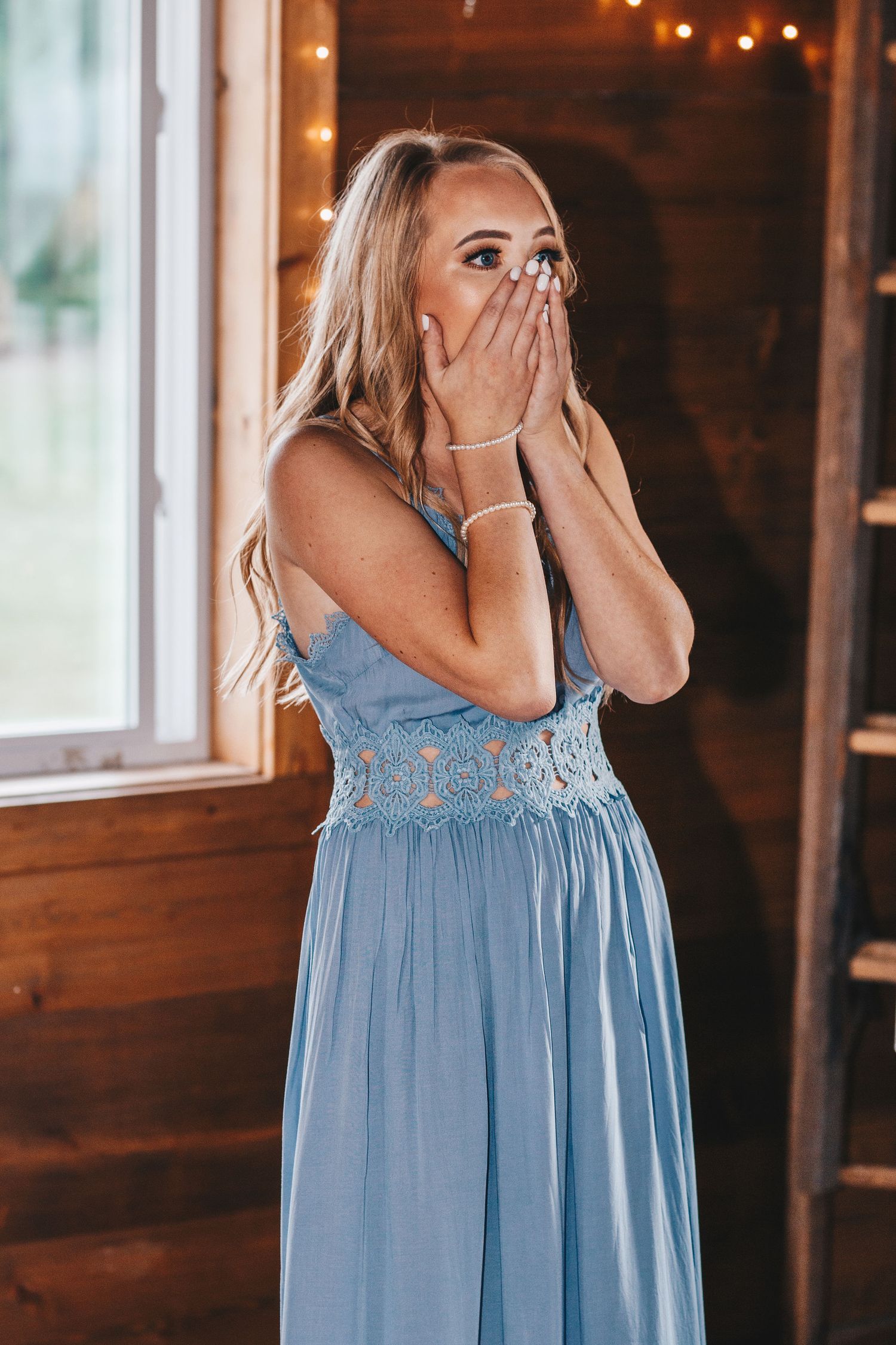 Surprised blonde woman in a blue lace dress covering her mouth with both hands indoors.