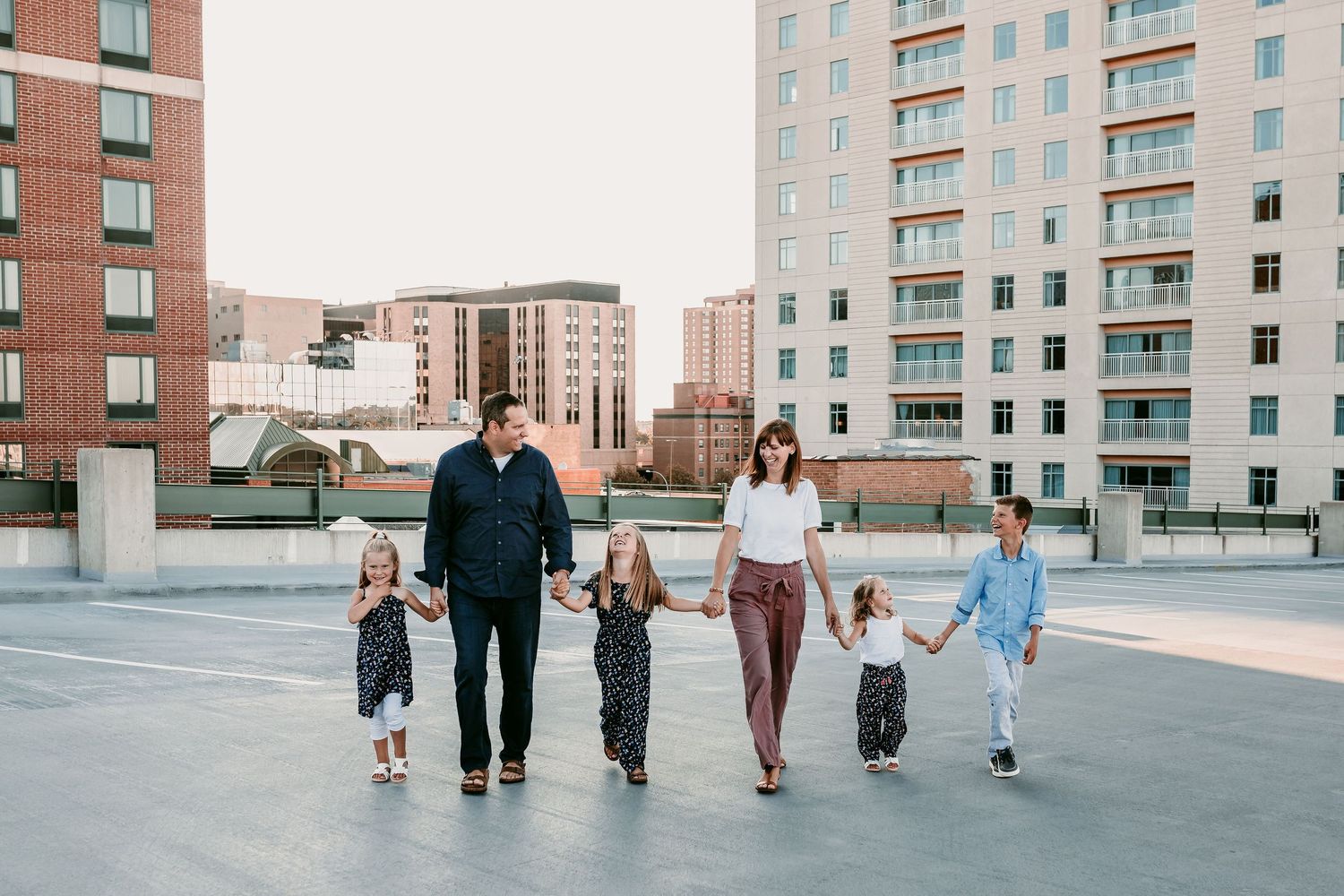 Happy family of six walking hand in hand across rooftop parking deck with city skyline in background.