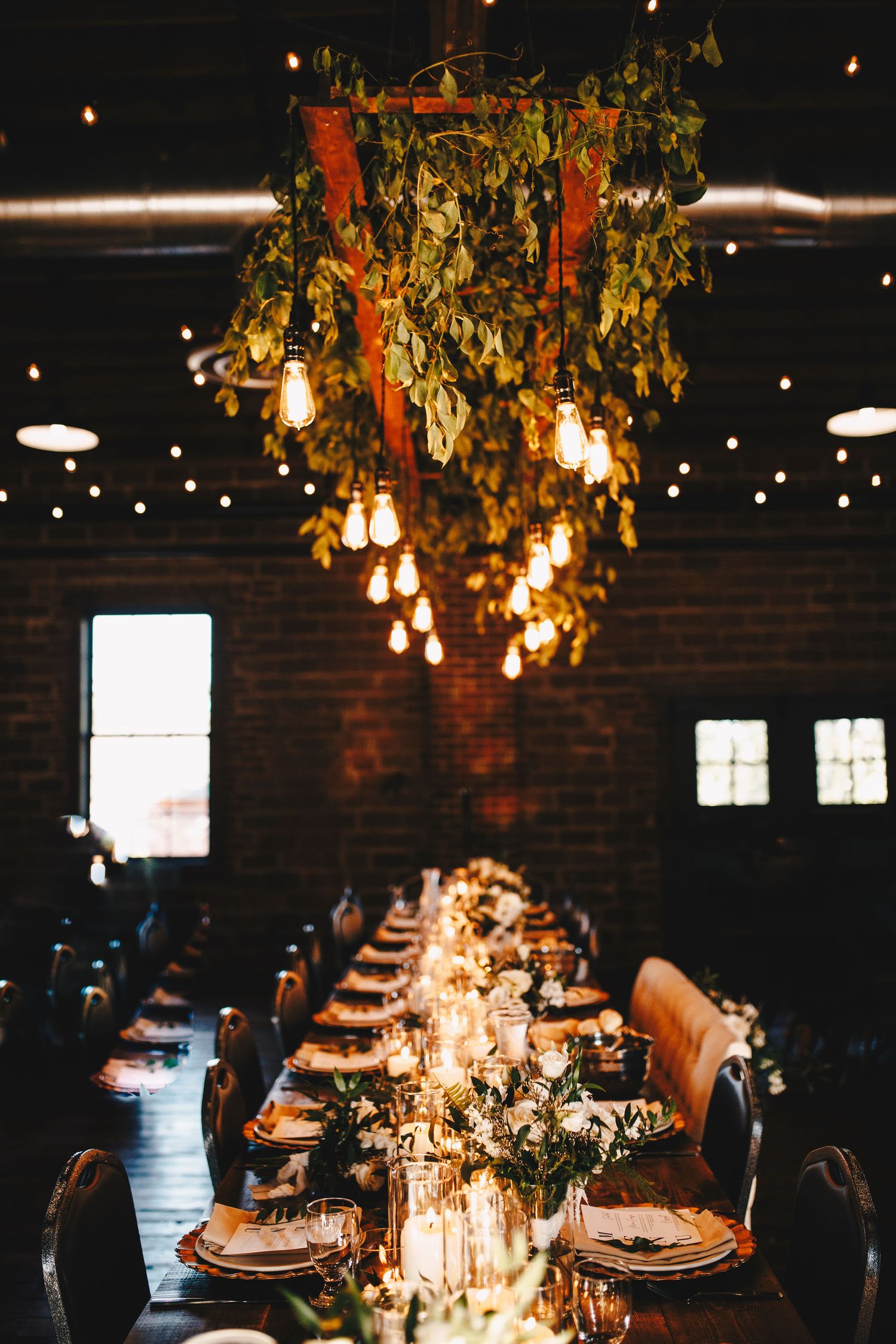 Elegant wedding reception hall with greenery chandelier, Edison bulbs, and long candlelit dining table.