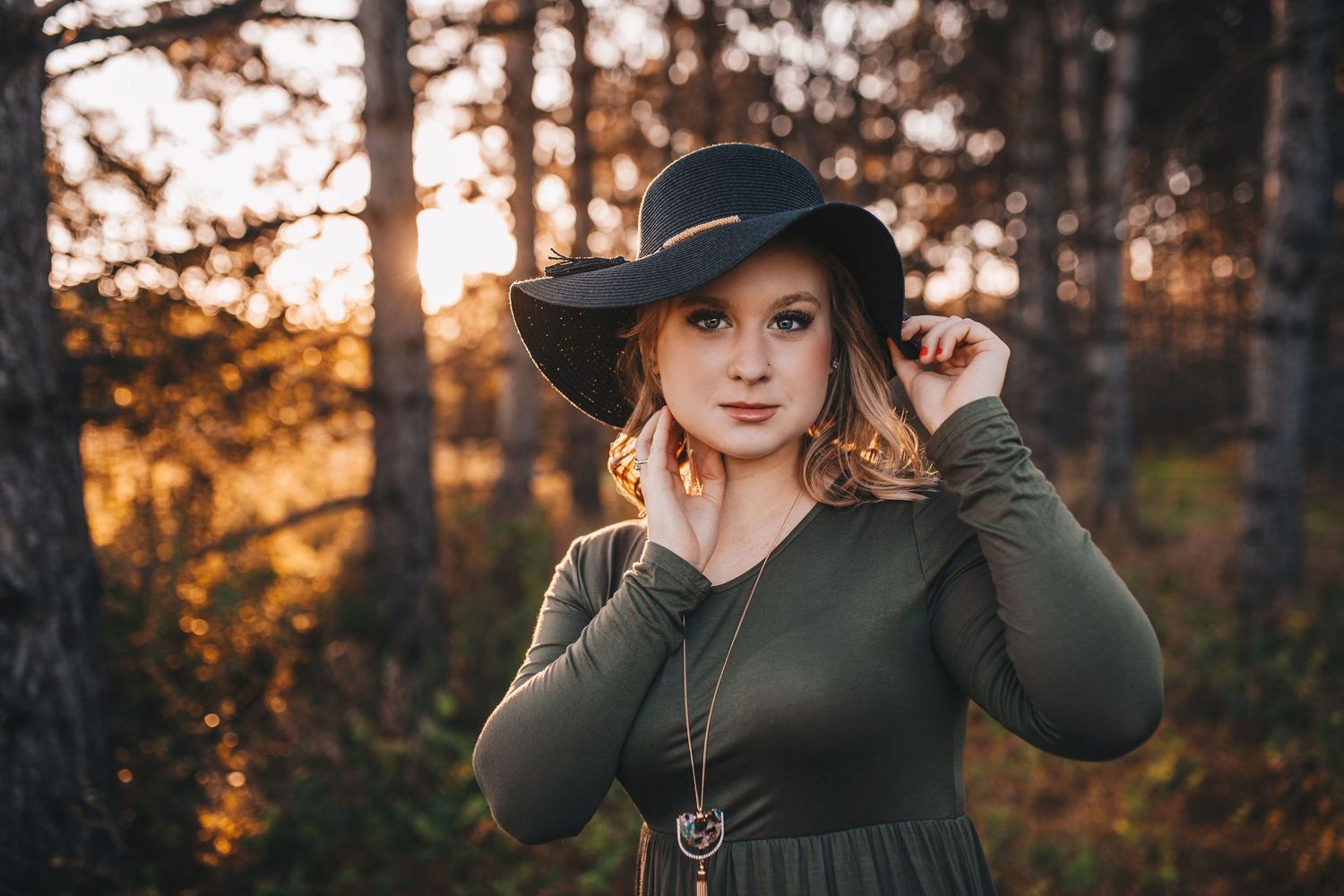 Young woman in green dress and wide-brim hat posing in autumn forest at sunset.