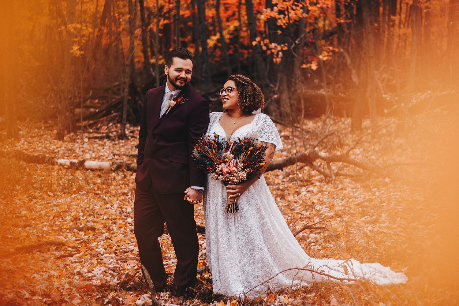 Bride in white lace gown surrounded by bridesmaids in rich burgundy and rust velvet dresses amid fall foliage.