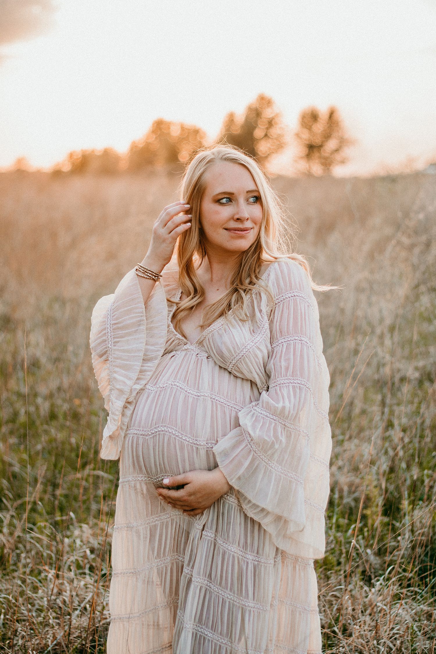 Pregnant woman in white boho dress holding belly in golden field at sunset.
