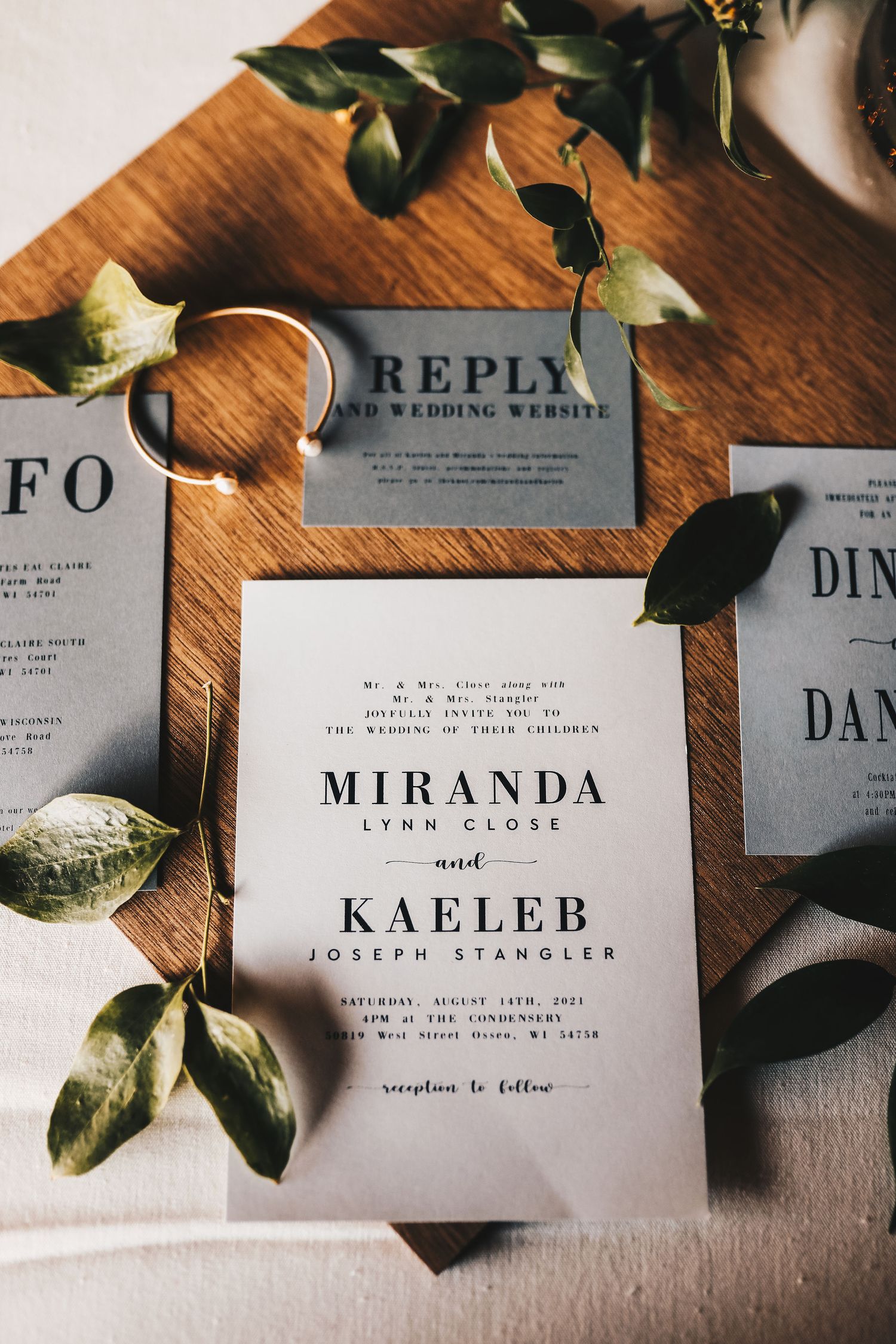 Elegant wedding invitation suite for Miranda Lynn Close and Kaeleb Joseph Stangler on wooden board with greenery.