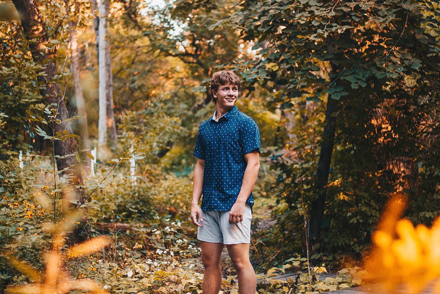 Smiling young man in blue shirt and shorts standing in colorful autumn forest.