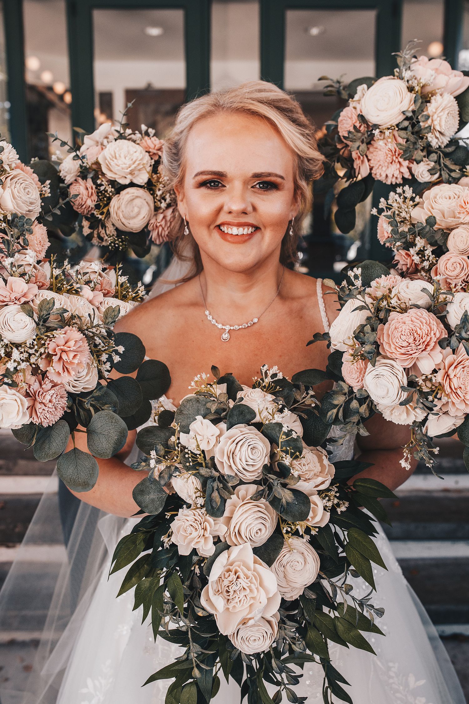 Smiling bride holding multiple bouquets of blush pink and white roses with eucalyptus greenery on her wedding day.