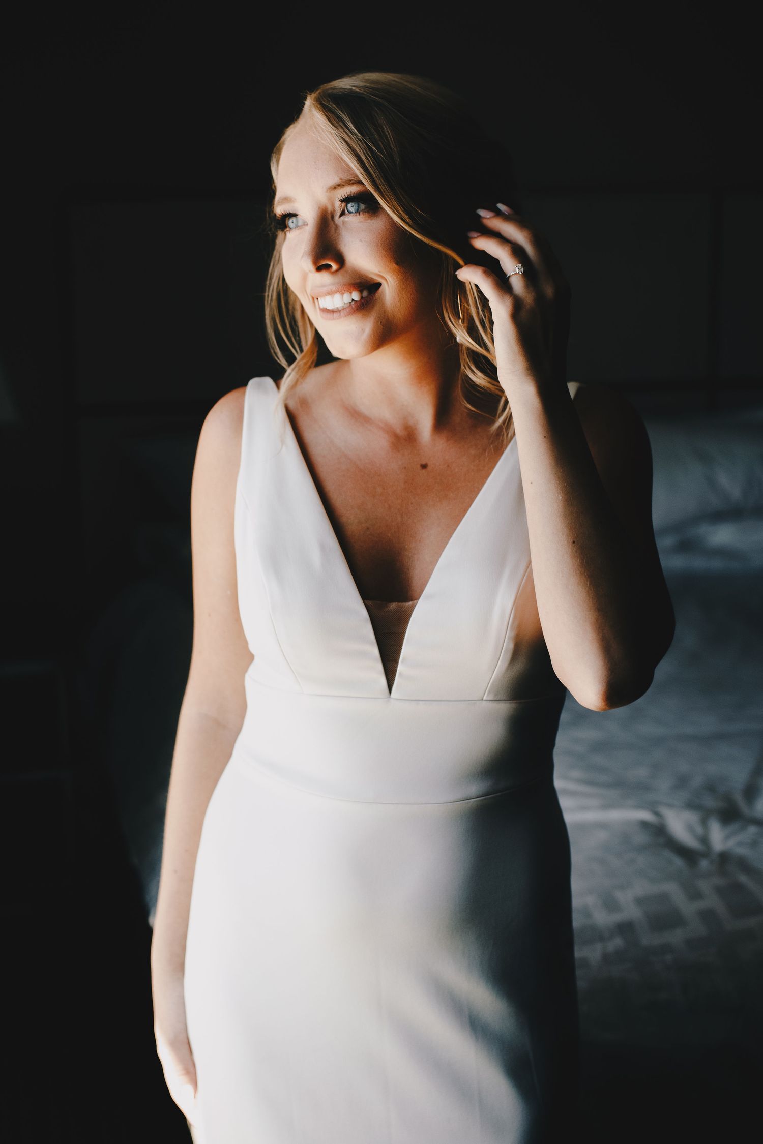 Radiant bride in sleek deep V-neck white wedding gown smiling in dramatic natural window light.