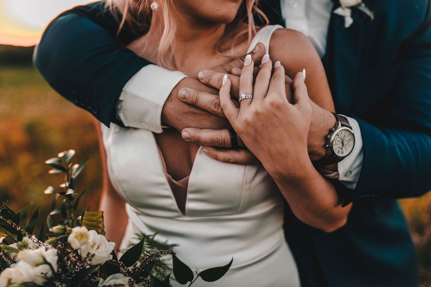 Bride and groom hold hands showing wedding rings while embracing at sunset.