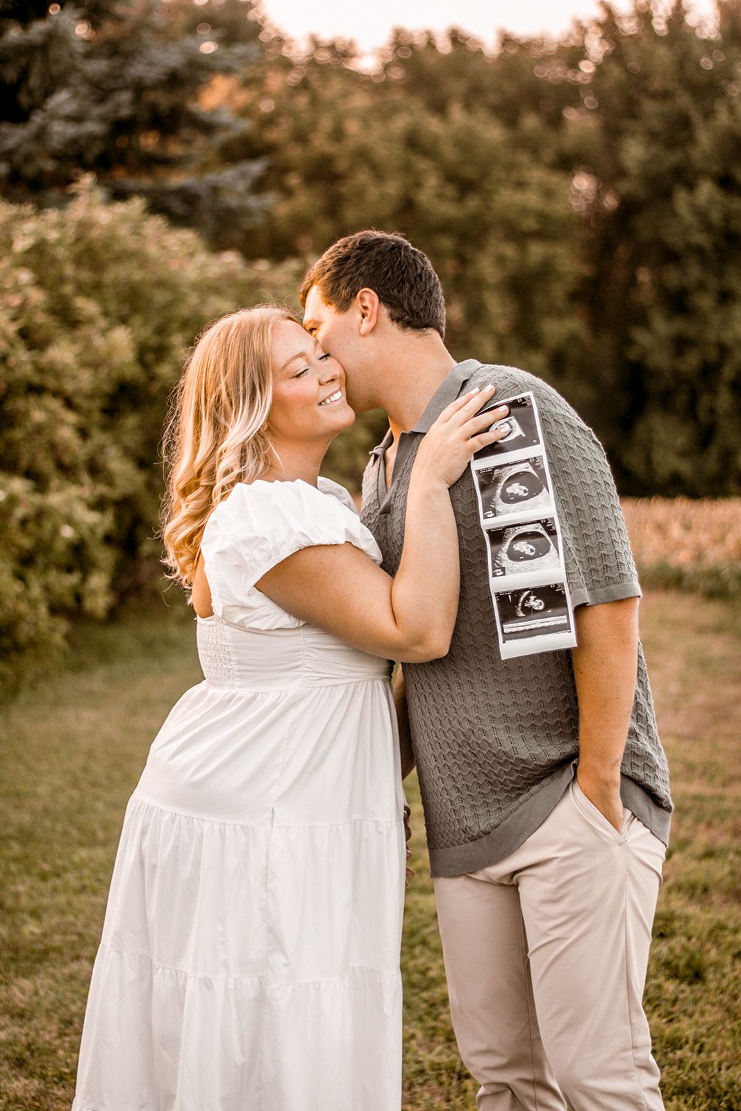Couple sharing a kiss outdoors holding ultrasound photos for a pregnancy announcement.