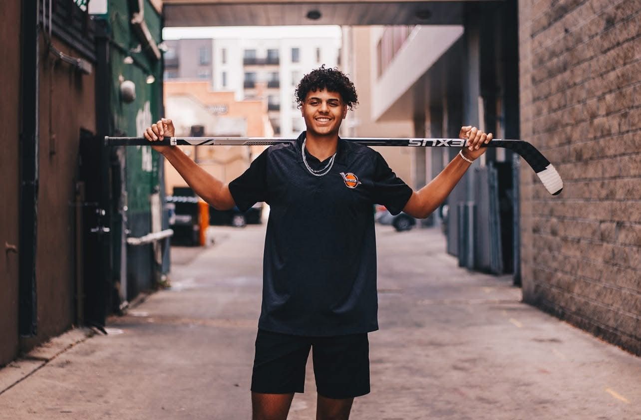 Smiling young man holding hockey stick across shoulders in urban alley.