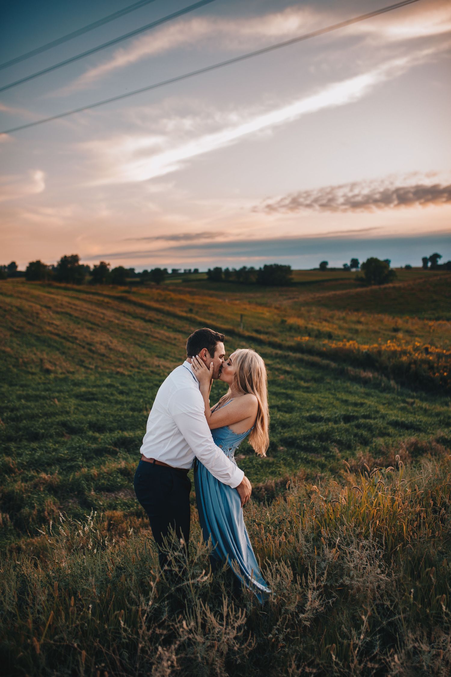 Couple shares passionate kiss in open green field during stunning golden sunset engagement photo session.