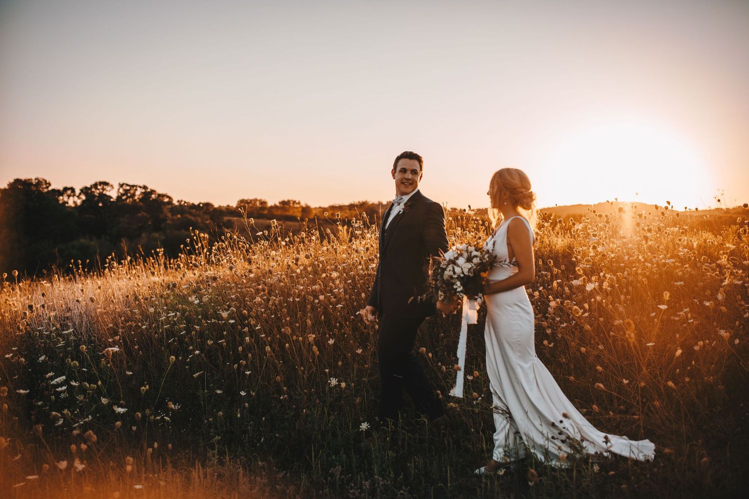 Bride and groom stand in a wildflower field holding a bouquet at golden hour.