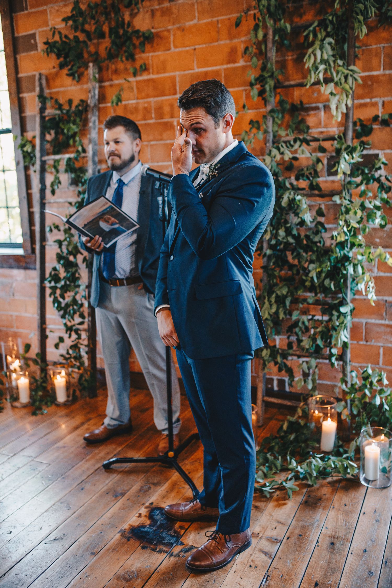 Emotional groom in navy suit wiping tears at altar decorated with greenery and candles.