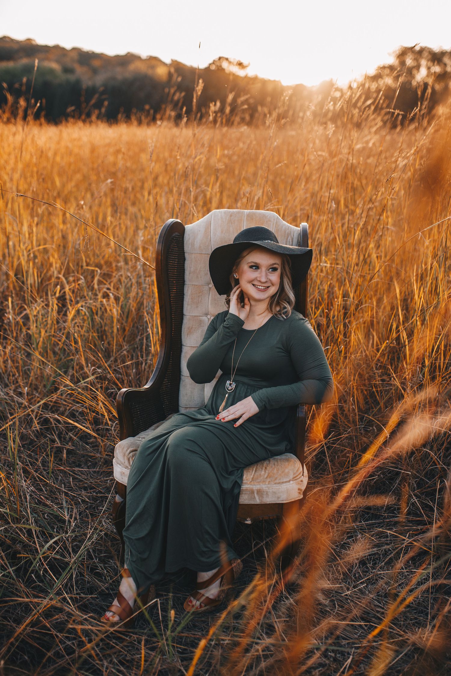 Smiling woman in green dress and black hat sitting in vintage chair in golden field.