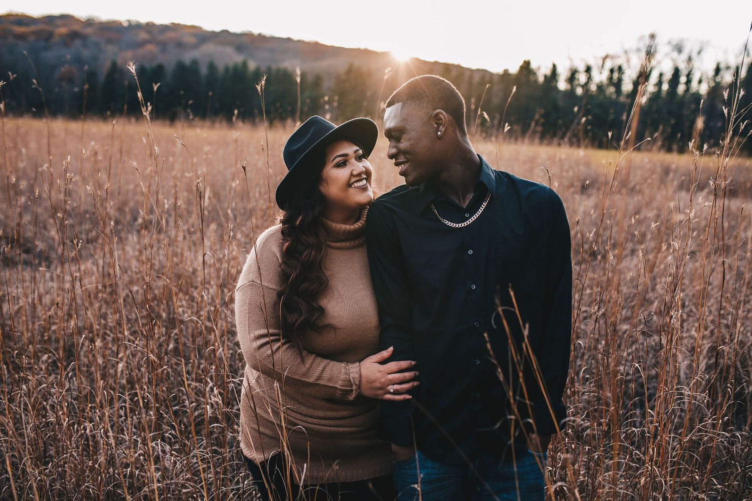 Couple smiles at each other in a golden field at sunset during an engagement session.