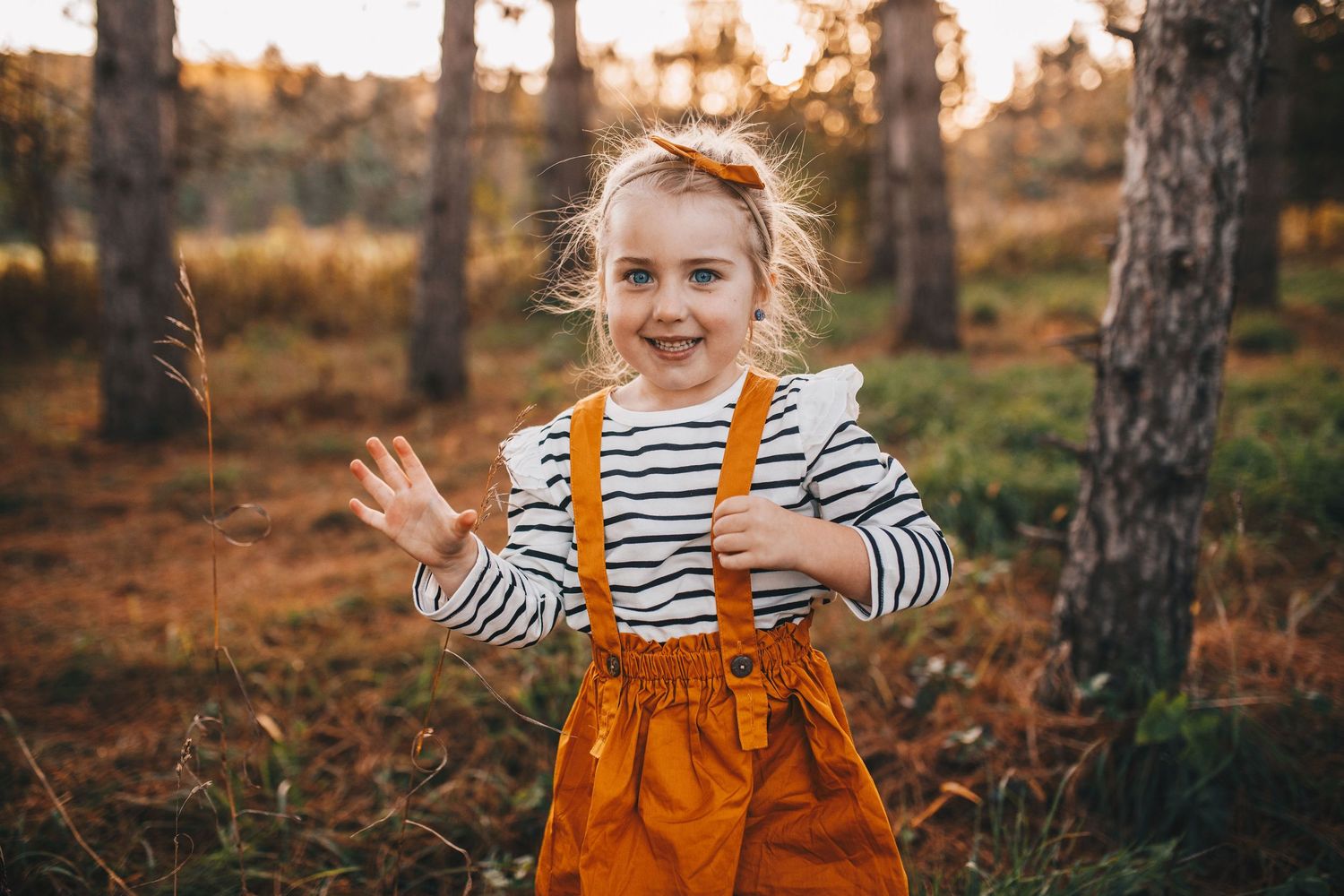 Joyful little girl in orange suspender skirt playing in autumn forest.