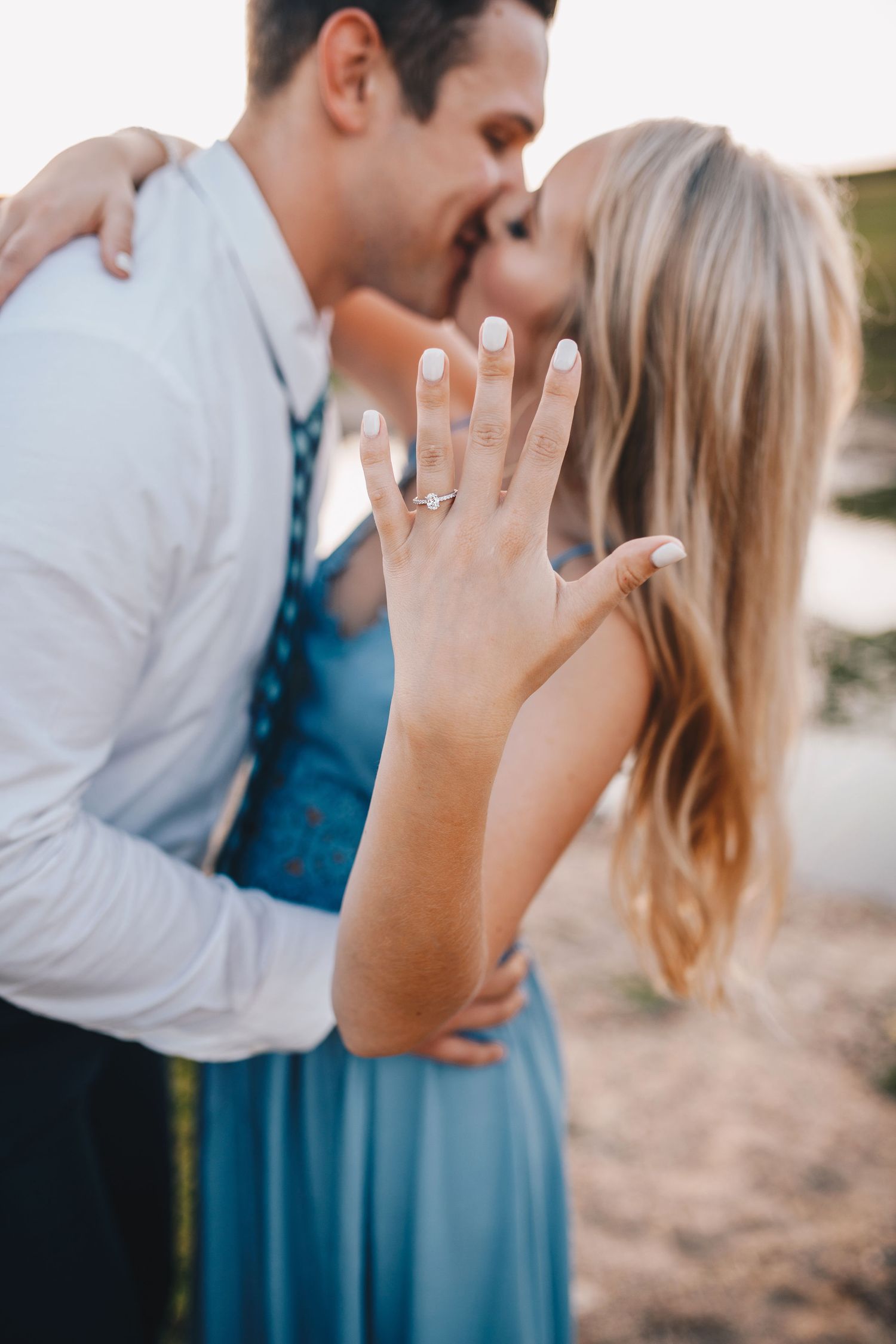 Newly engaged woman displays diamond ring while kissing partner outdoors in soft golden light.
