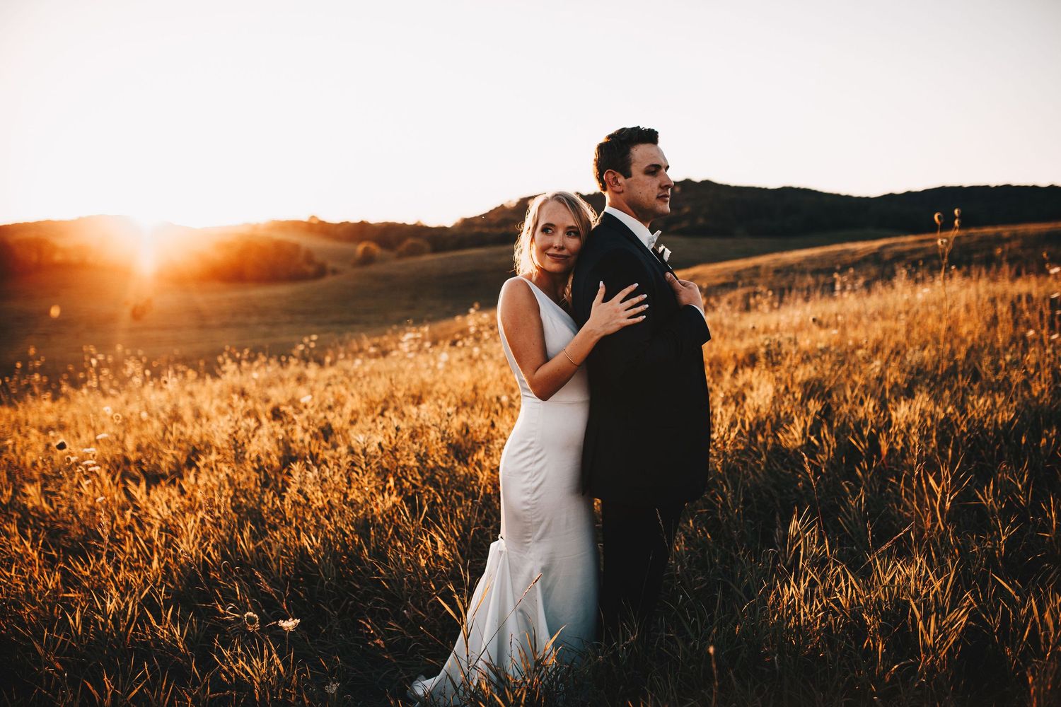 Bride leans against groom in a golden meadow during a stunning sunset wedding shoot.
