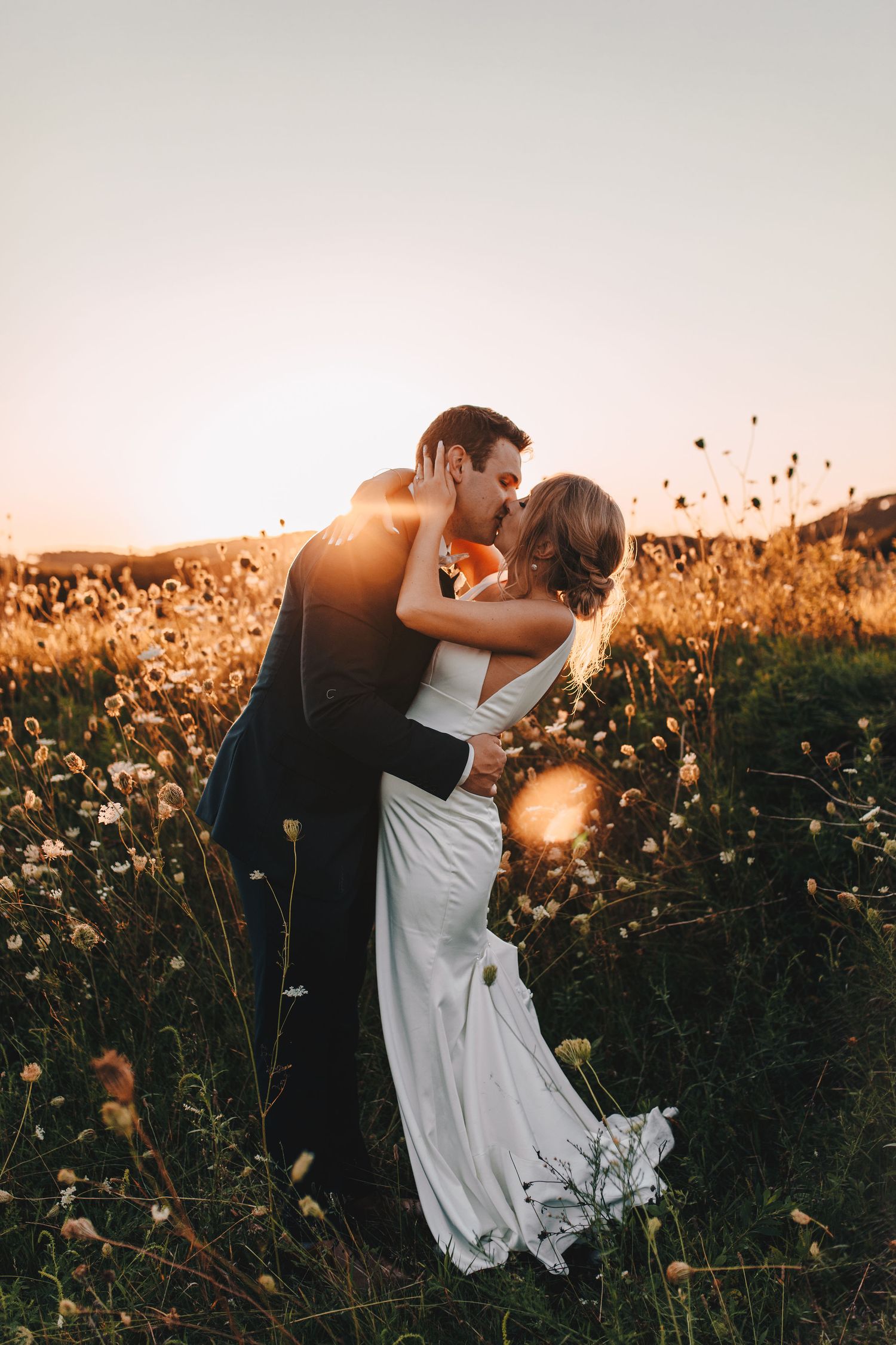 Bride and groom share a passionate kiss in a wildflower field at sunset.