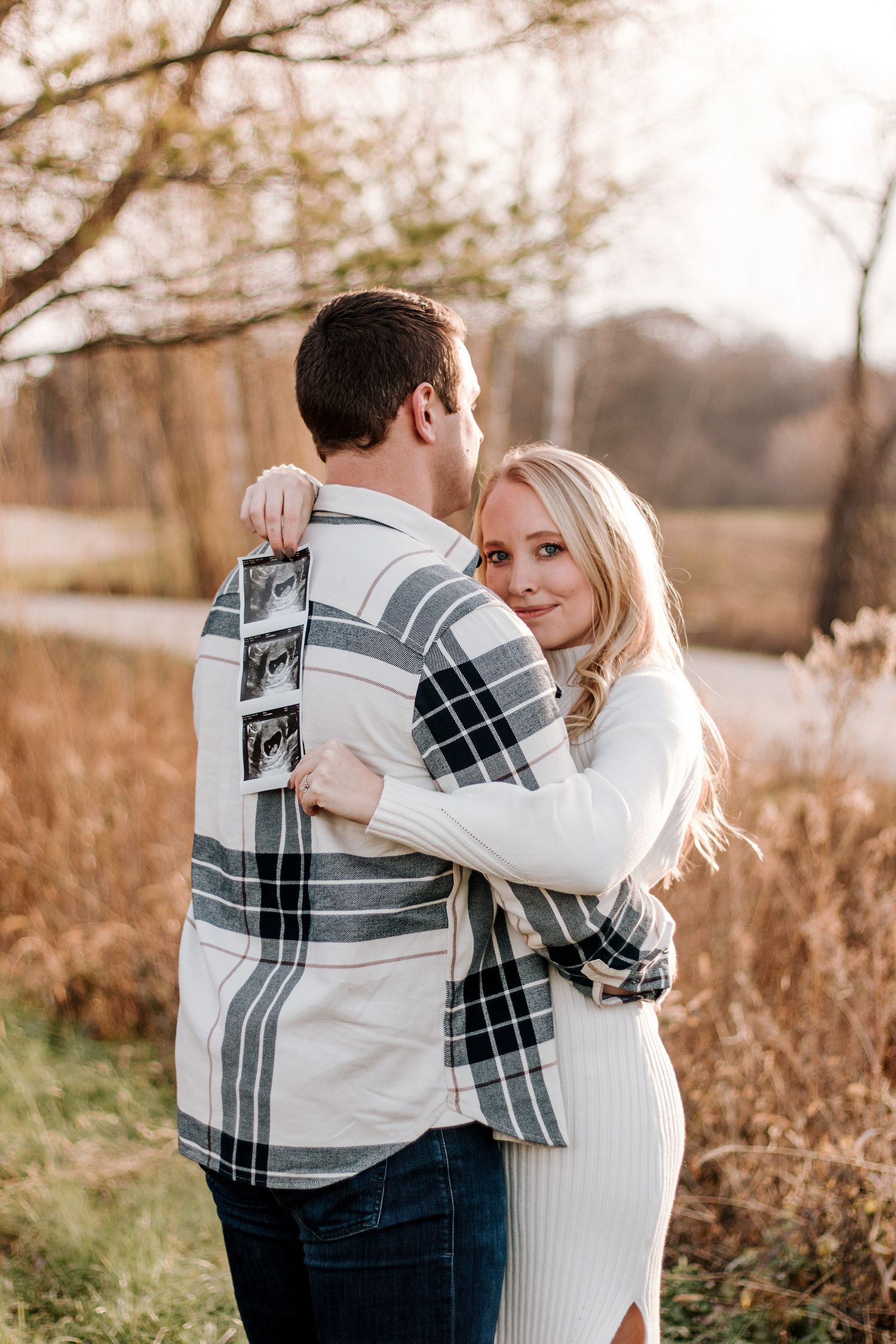 Couple embracing outdoors with ultrasound photos for a fall pregnancy announcement.