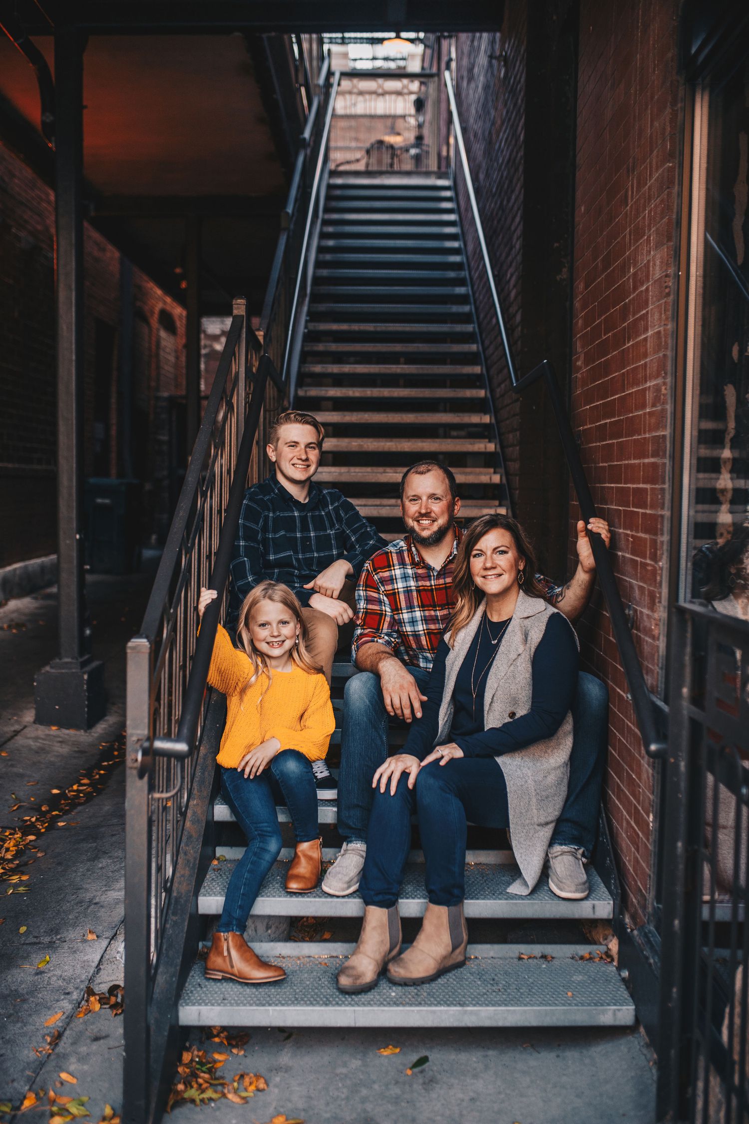Family of four posing on urban metal staircase with brick walls, wearing coordinated fall outfits in warm tones.