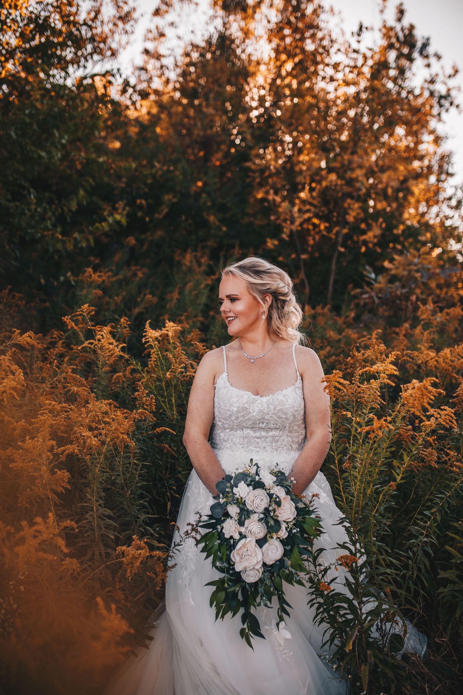 Bride in white lace gown holding bouquet surrounded by golden autumn wildflowers at sunset.