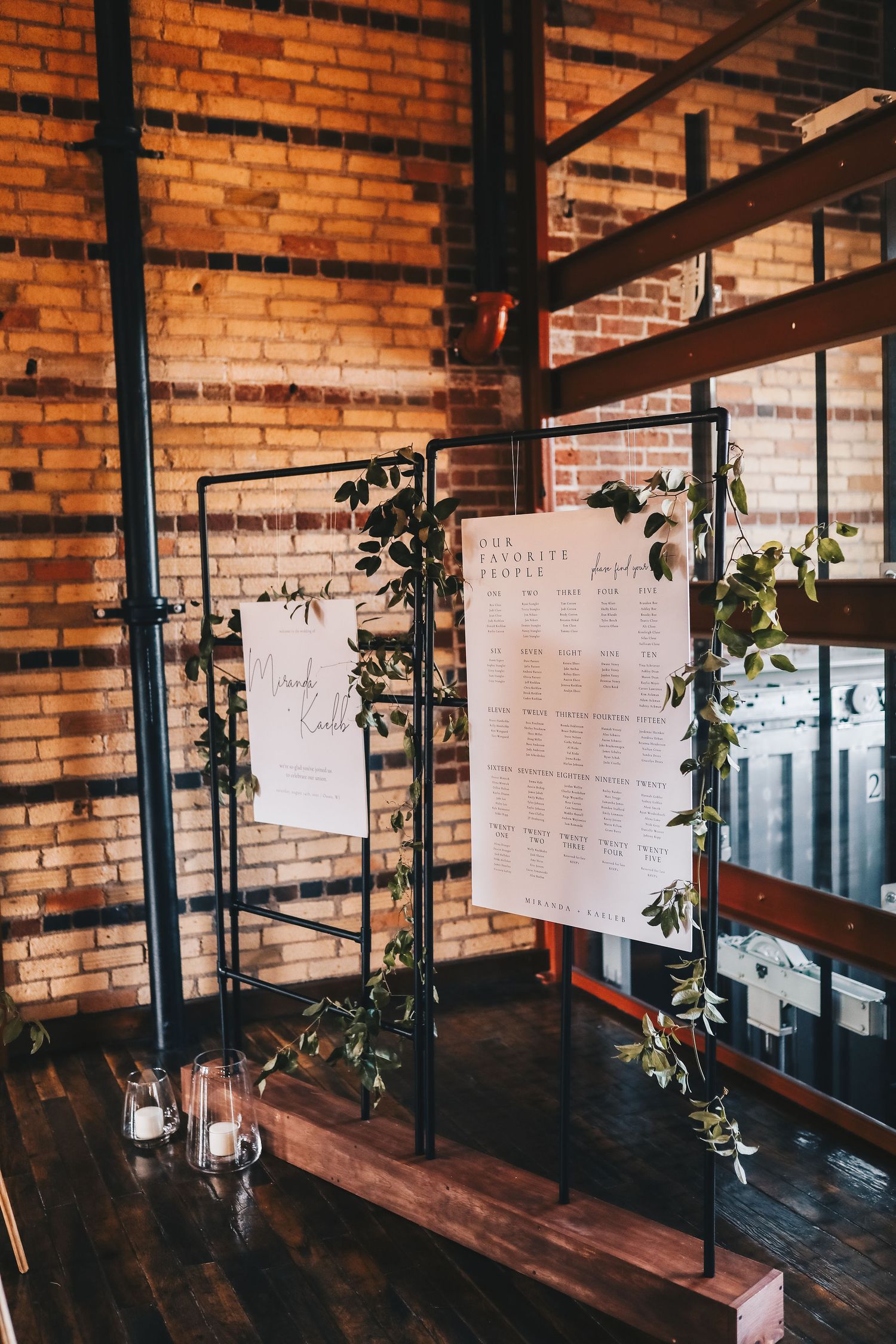 Industrial wedding seating chart display on black metal shelving with greenery against exposed brick wall.