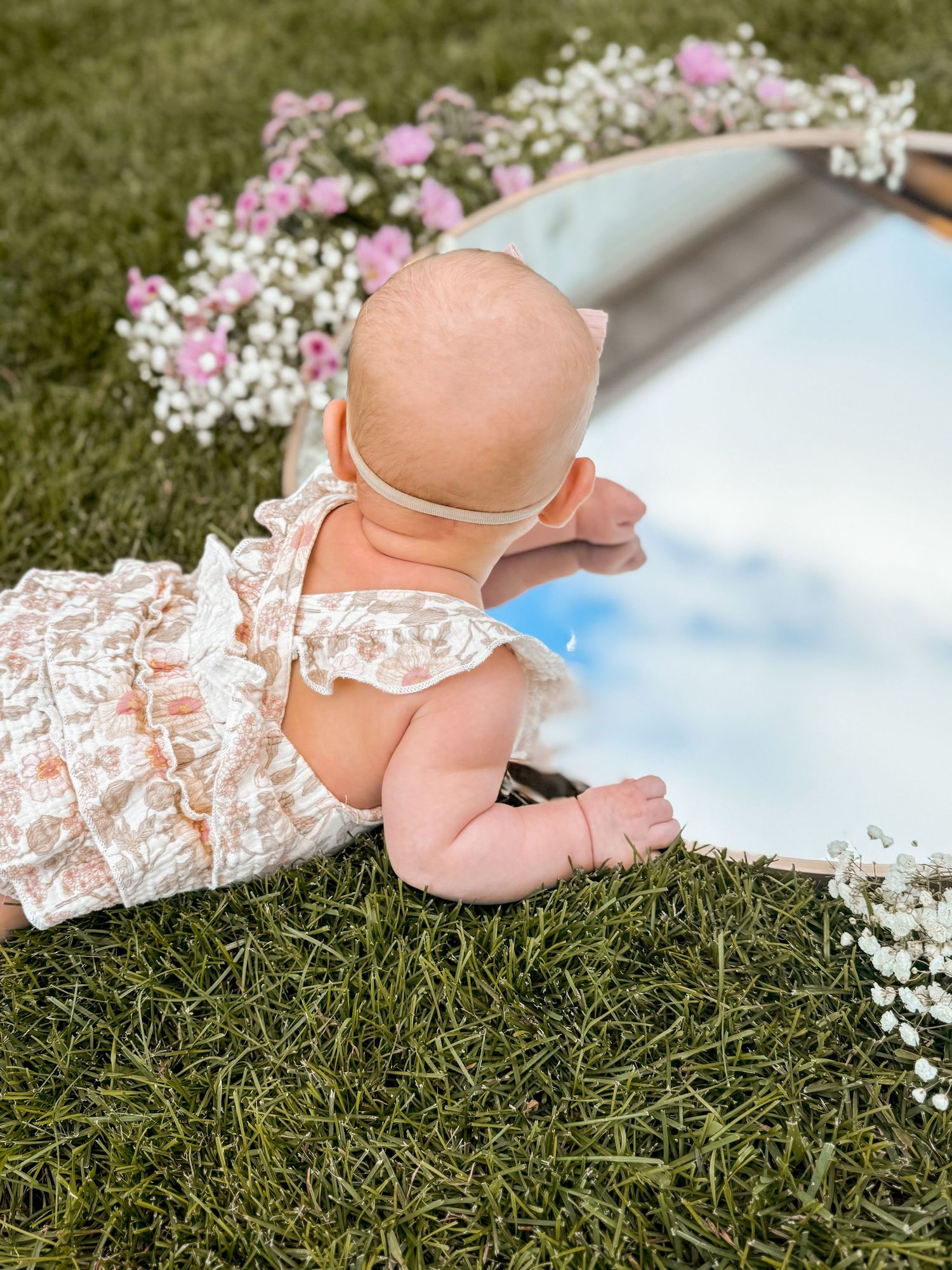 Baby in floral outfit lying on grass near mirror surrounded by pink flowers.