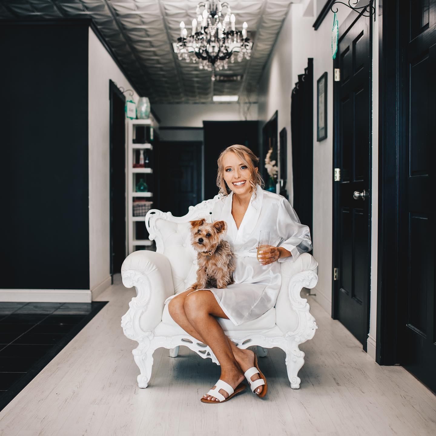 Bride in white robe sitting on ornate white chair holding Yorkshire Terrier in salon.