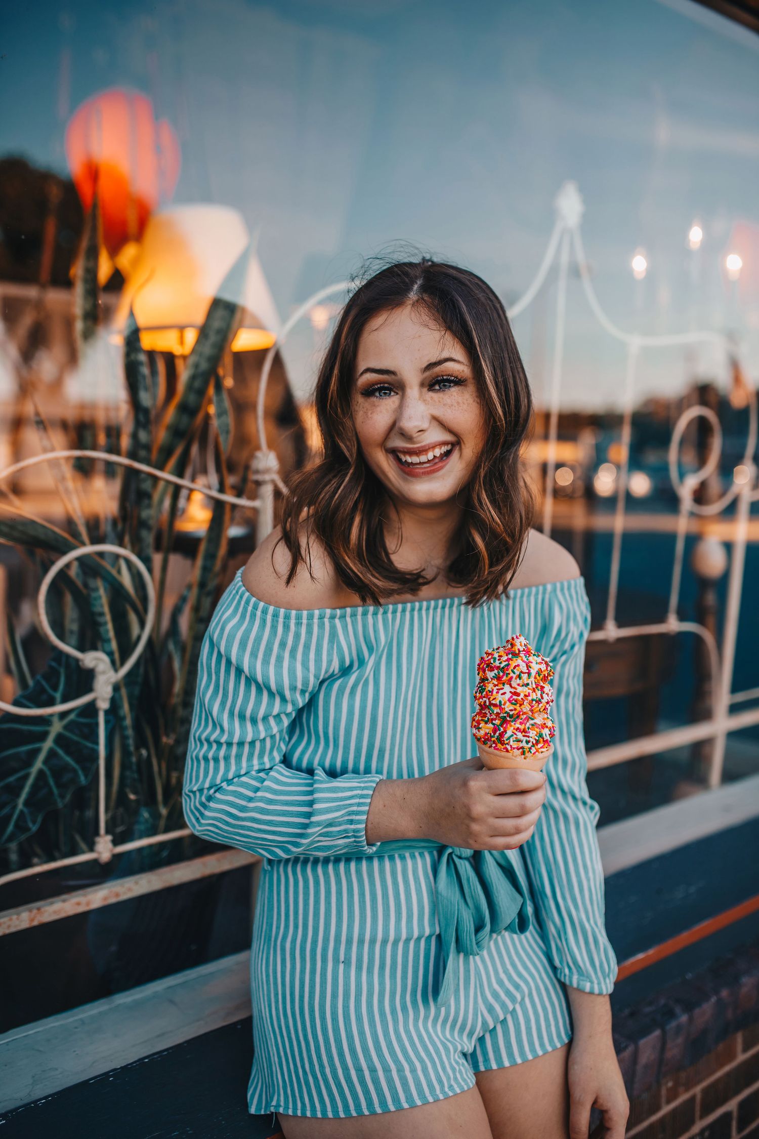 Happy brunette woman in striped off-shoulder romper holding a sprinkle-covered ice cream cone.
