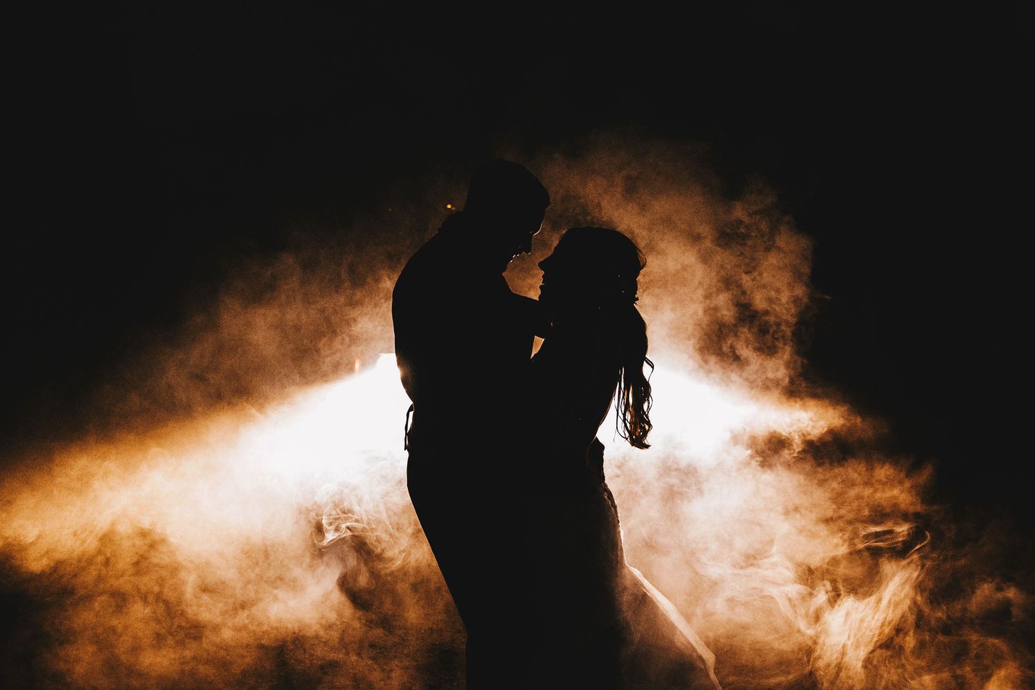 Silhouette of bride and groom embracing in dramatic smoke and golden backlight.