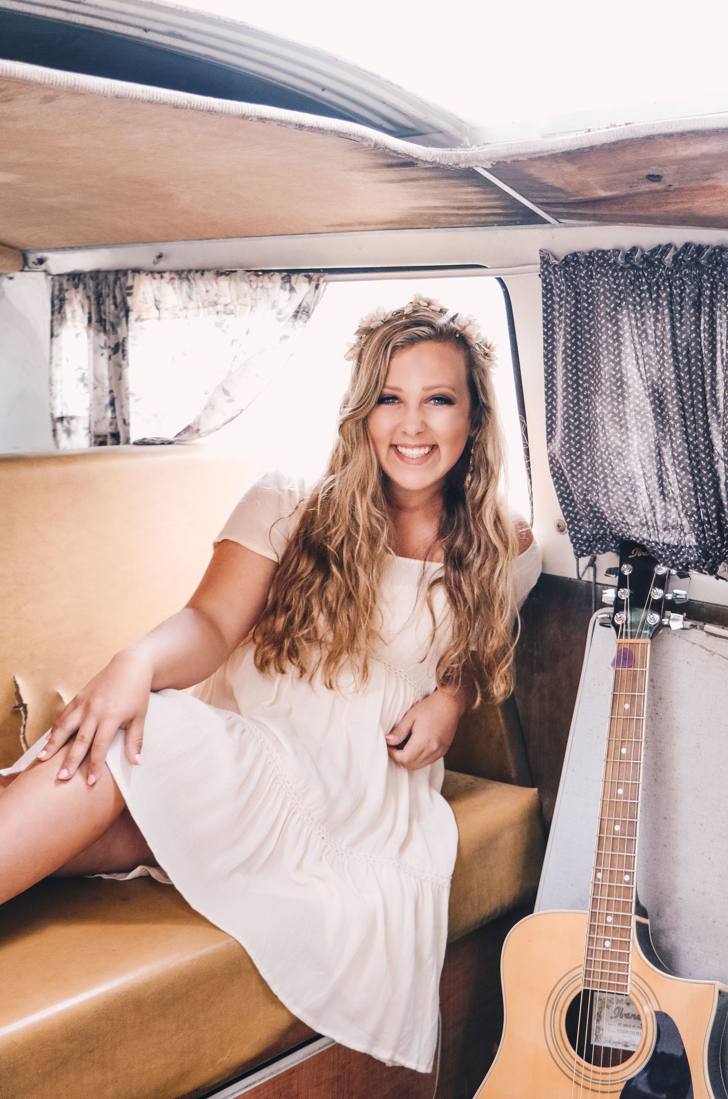 Smiling young woman in white dress sitting in vintage van with acoustic guitar.