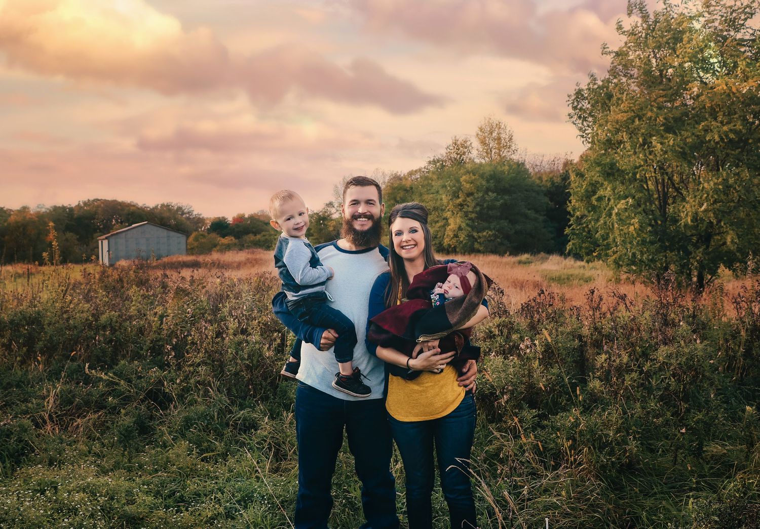 Young family of four posing in a field at sunset with dramatic pink sky.