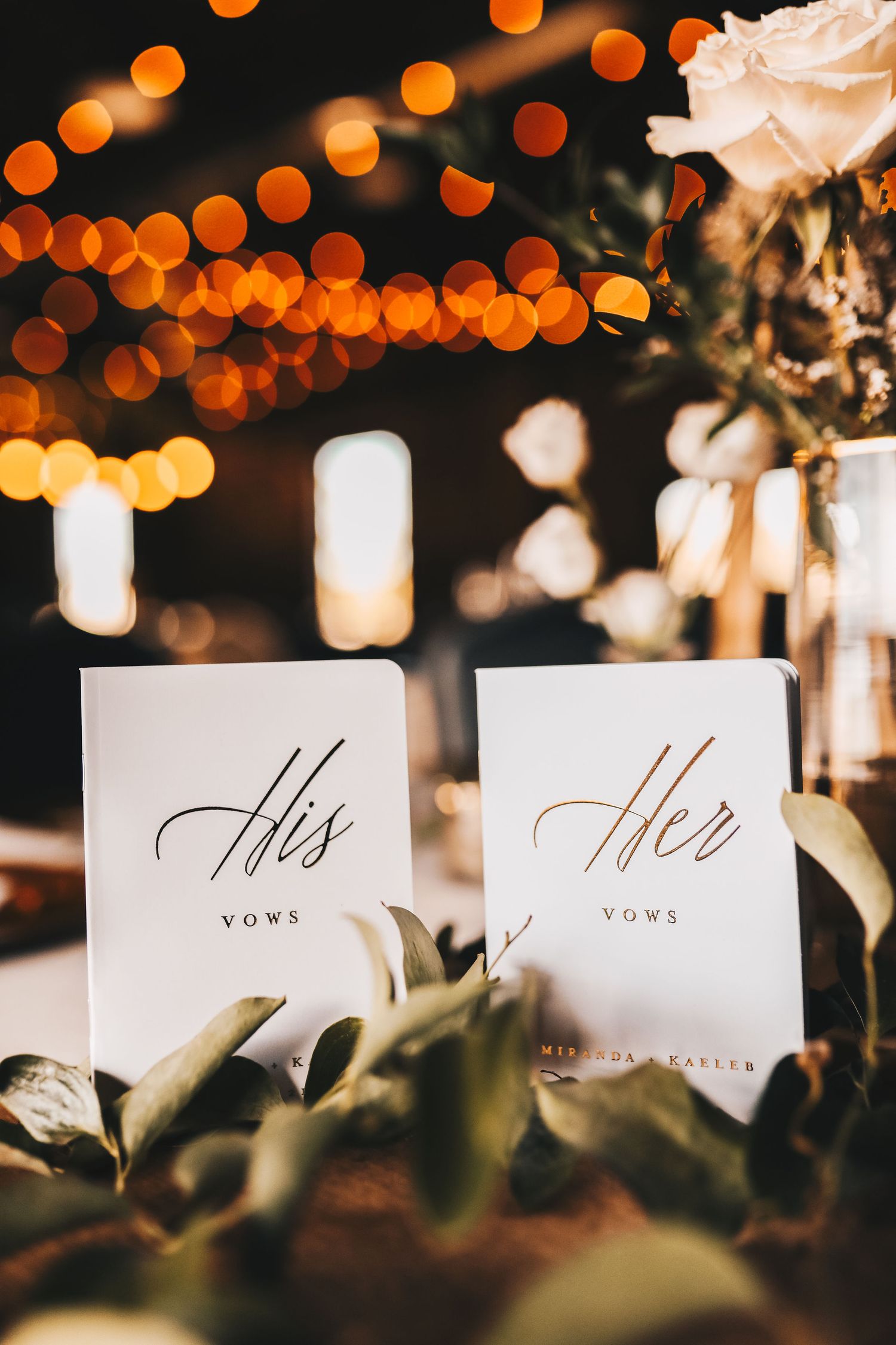 Elegant His and Her vows booklets displayed among greenery with warm bokeh lights background.