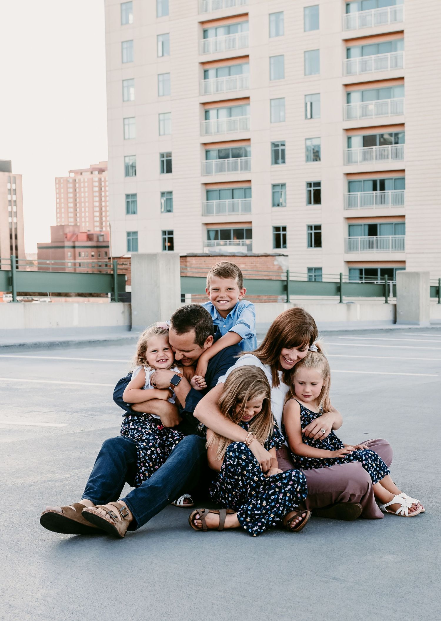Joyful family of six piled together laughing on urban rooftop parking deck with city buildings behind them.