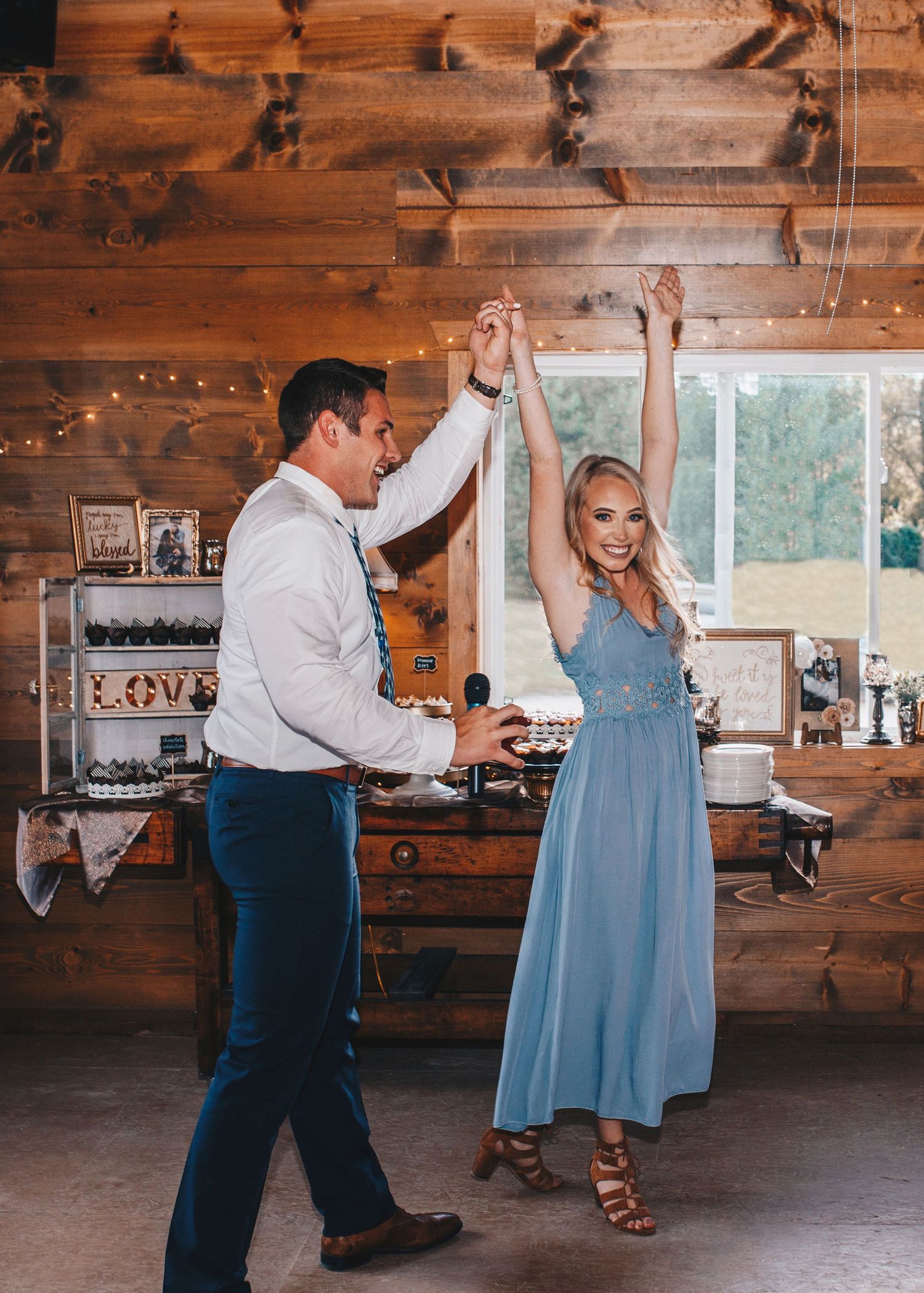 Couple dancing joyfully in a rustic wooden barn venue decorated with string lights.