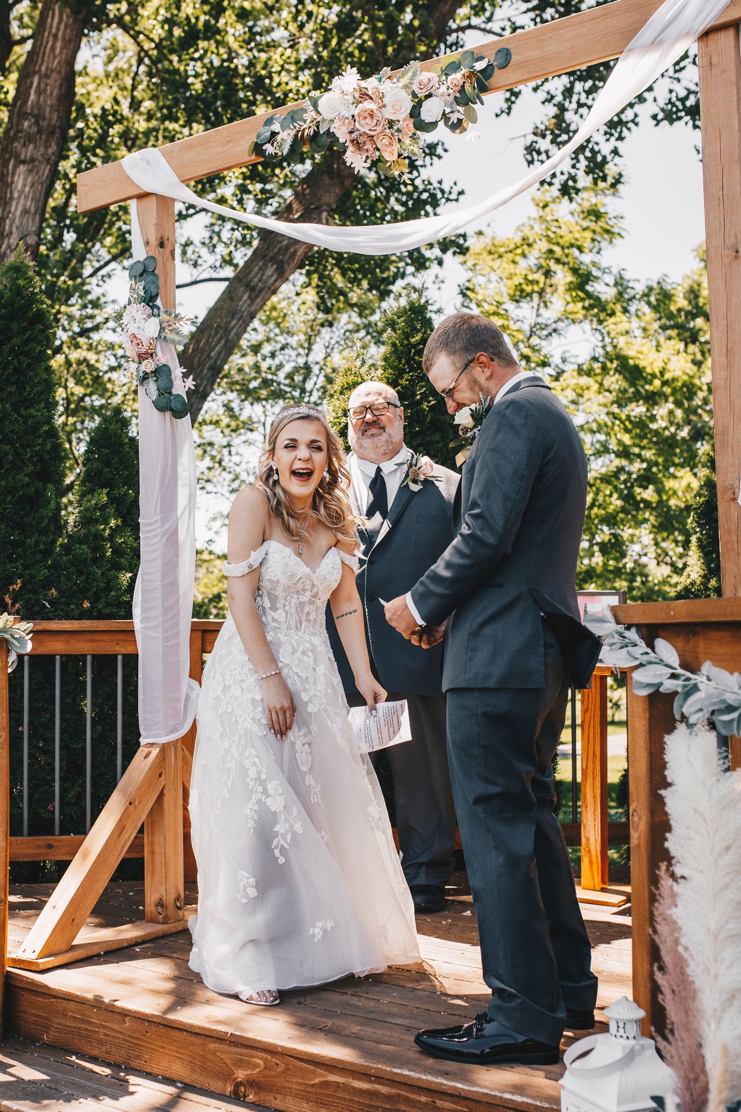 Bride laughing joyfully with groom during outdoor wedding ceremony under wooden floral arch.