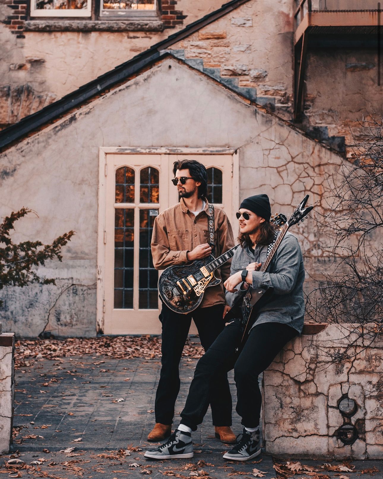 Two guitarists posing with electric guitars outside a vintage stone building in autumn.